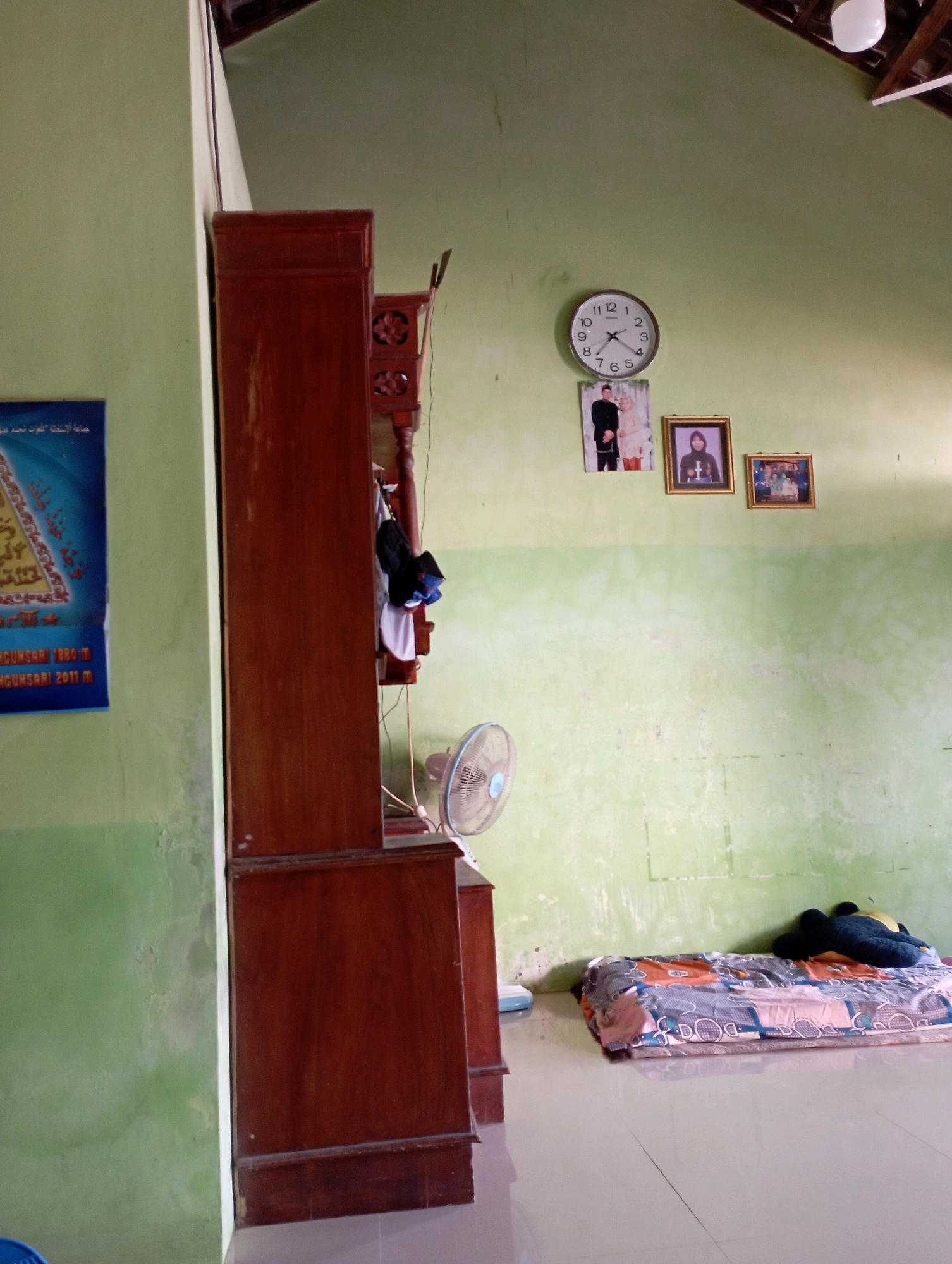 The image depicts a room with light green walls and a tiled floor, likely in Sonobekel, Indonesia. On the left, a tall, dark wooden cabinet stands against the wall, with some clothing items hanging from it. A framed poster is partially visible on the wall to the left of the cabinet. In the center of the room, a round wall clock with black numerals and hands is mounted on the wall above three framed photographs. The photographs show a couple, a person in a headscarf, and a group of people. To the right, a single bed with a patterned mattress is placed on the floor, with a dark stuffed toy lying on the pillow. A small, round fan is positioned in front of the cabinet, near the floor. The ceiling is visible at the top, with exposed wooden beams and a light fixture. The lighting suggests it is daytime, possibly with natural light entering the room.