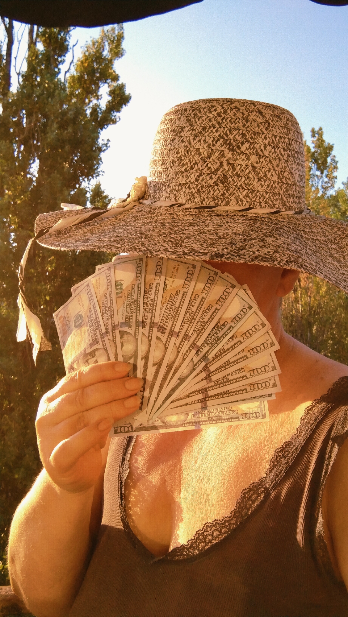 The image shows a person wearing a wide-brimmed hat and a dark brown tank top with lace trim. The person is holding a fan of US dollar bills in their right hand, obscuring their face. The background consists of trees and a clear blue sky, suggesting an outdoor setting. The lighting indicates it is daytime, with the sun casting a warm glow on the scene. There is no discernible information in the image that would allow for a determination of the location in Horishni Plavni, Ukraine.