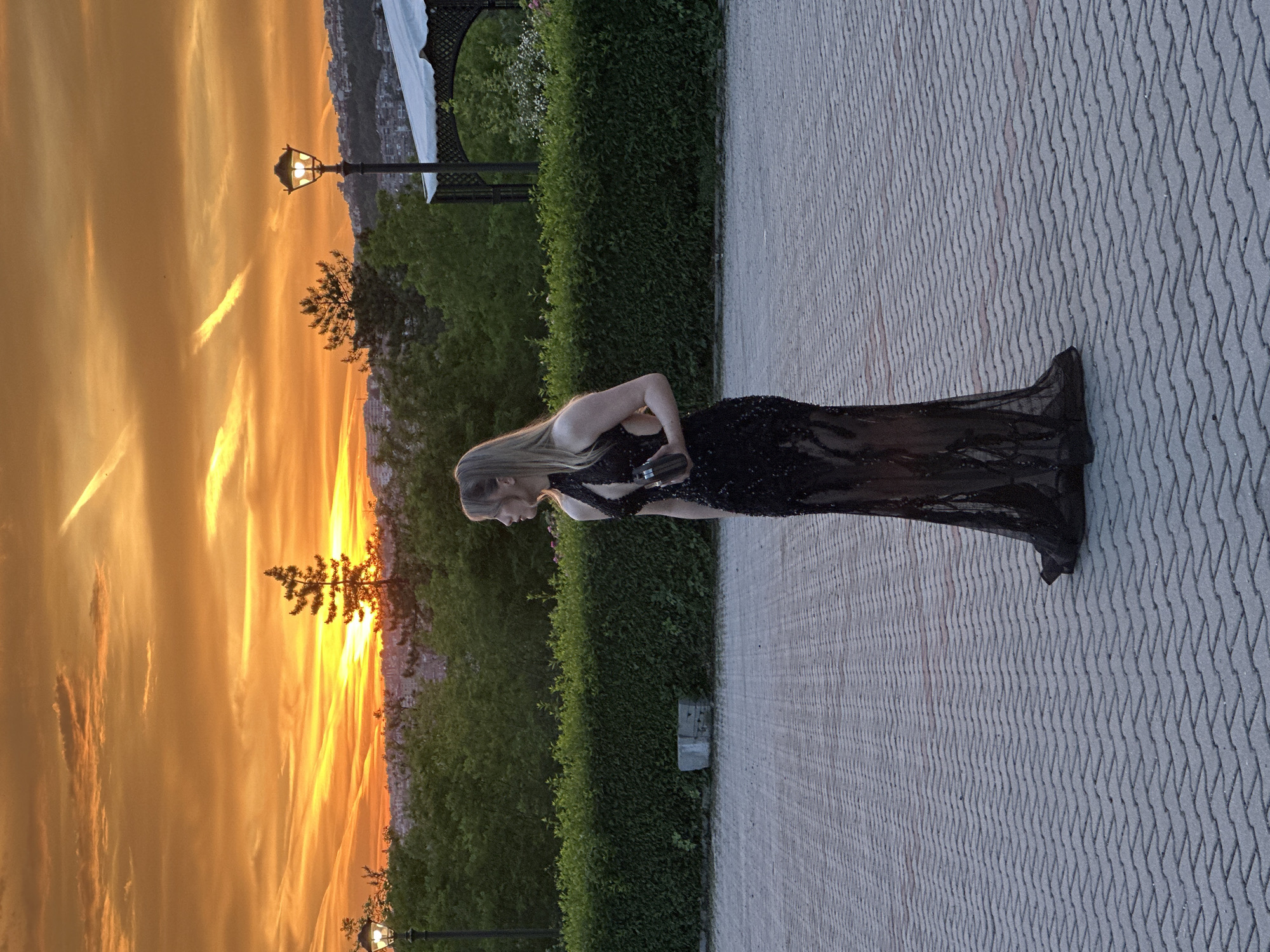 A woman in a black dress stands on a paved walkway next to a manicured hedge. The background reveals an orange sunset and a street lamp. This scene appears to be taken in Veliko Tarnovo, Bulgaria, specifically overlooking the city from above.
