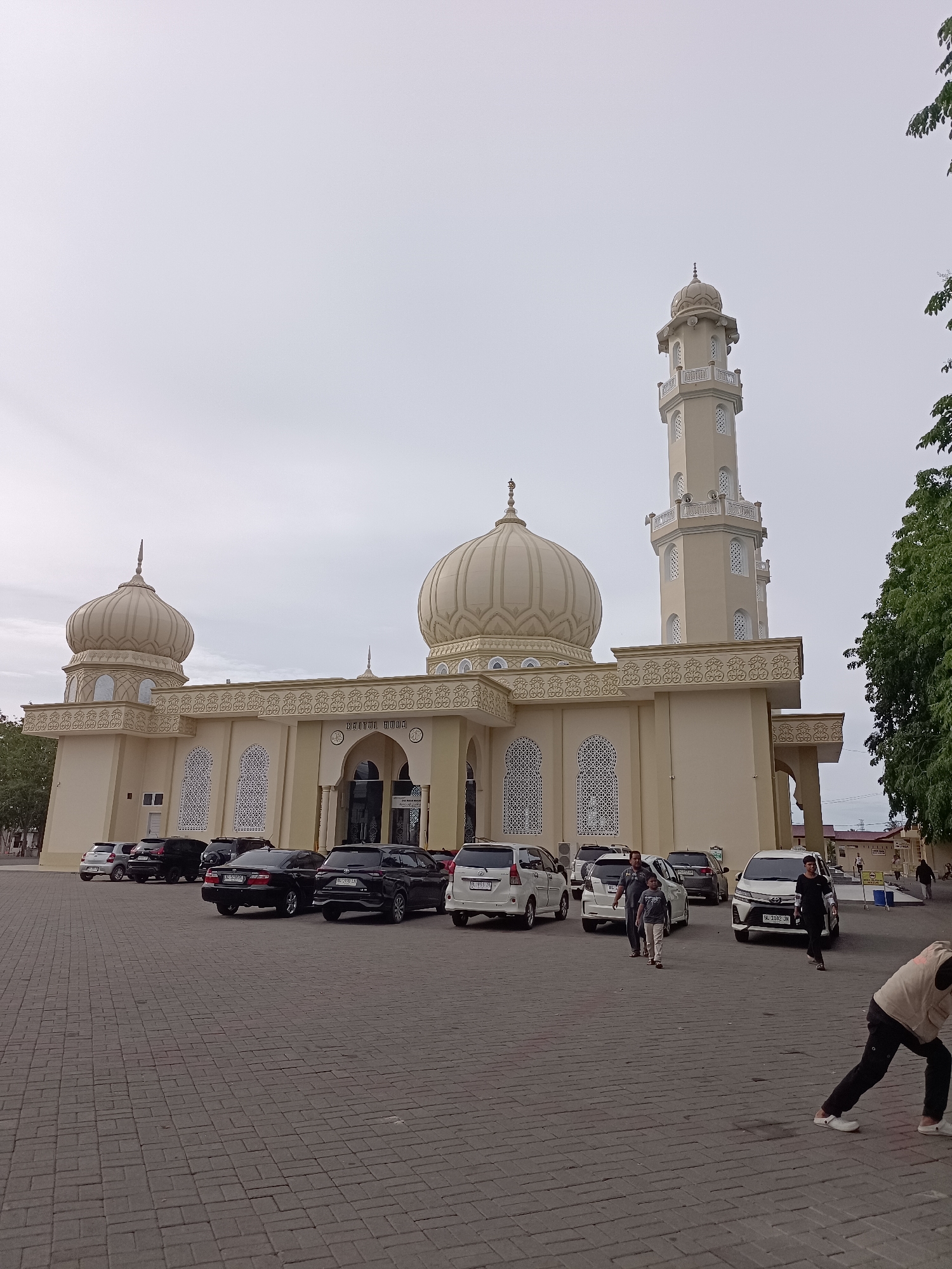 The image depicts the Baitul Huda Mosque in the city of Mee Tanjong Usi, Indonesia, under a cloudy sky. The mosque features a prominent minaret and several domes, with light-colored walls and decorative latticework on the windows. A paved courtyard in front of the mosque is occupied by several parked cars, including sedans and SUVs. People are visible in the courtyard, some walking and others standing near the cars. The overall scene suggests a public gathering or an active day at the mosque.