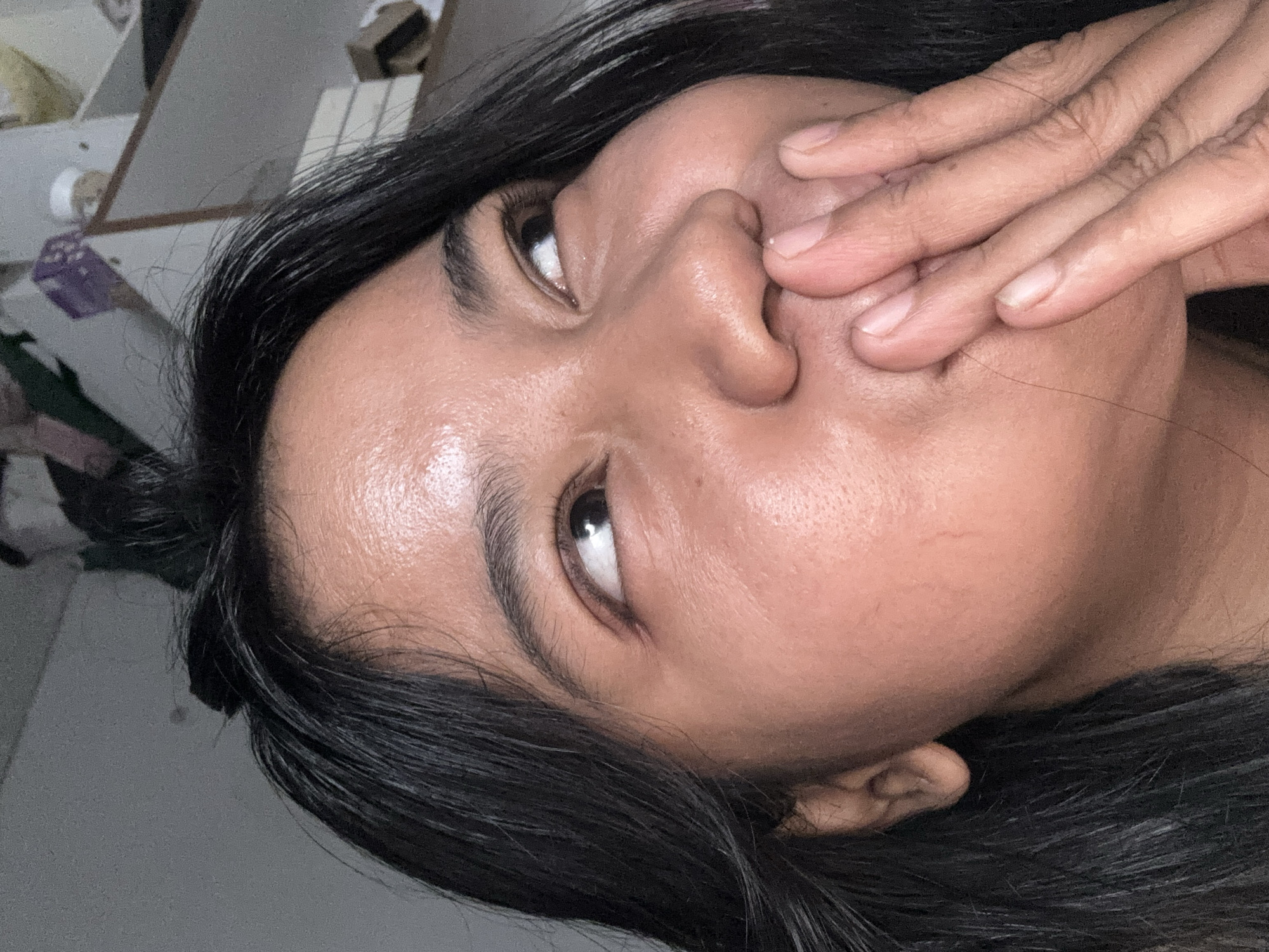 The image is a close-up portrait of a young woman with dark hair and medium-toned skin. She is looking up and to her right, with her hand covering her mouth. Her expression appears thoughtful or amused. The background is dimly lit and appears to be an indoor setting, possibly a room with shelves in the distance. The image does not contain any elements that allow for specific identification of its location within North Jakarta, Indonesia, or any other urban environment.