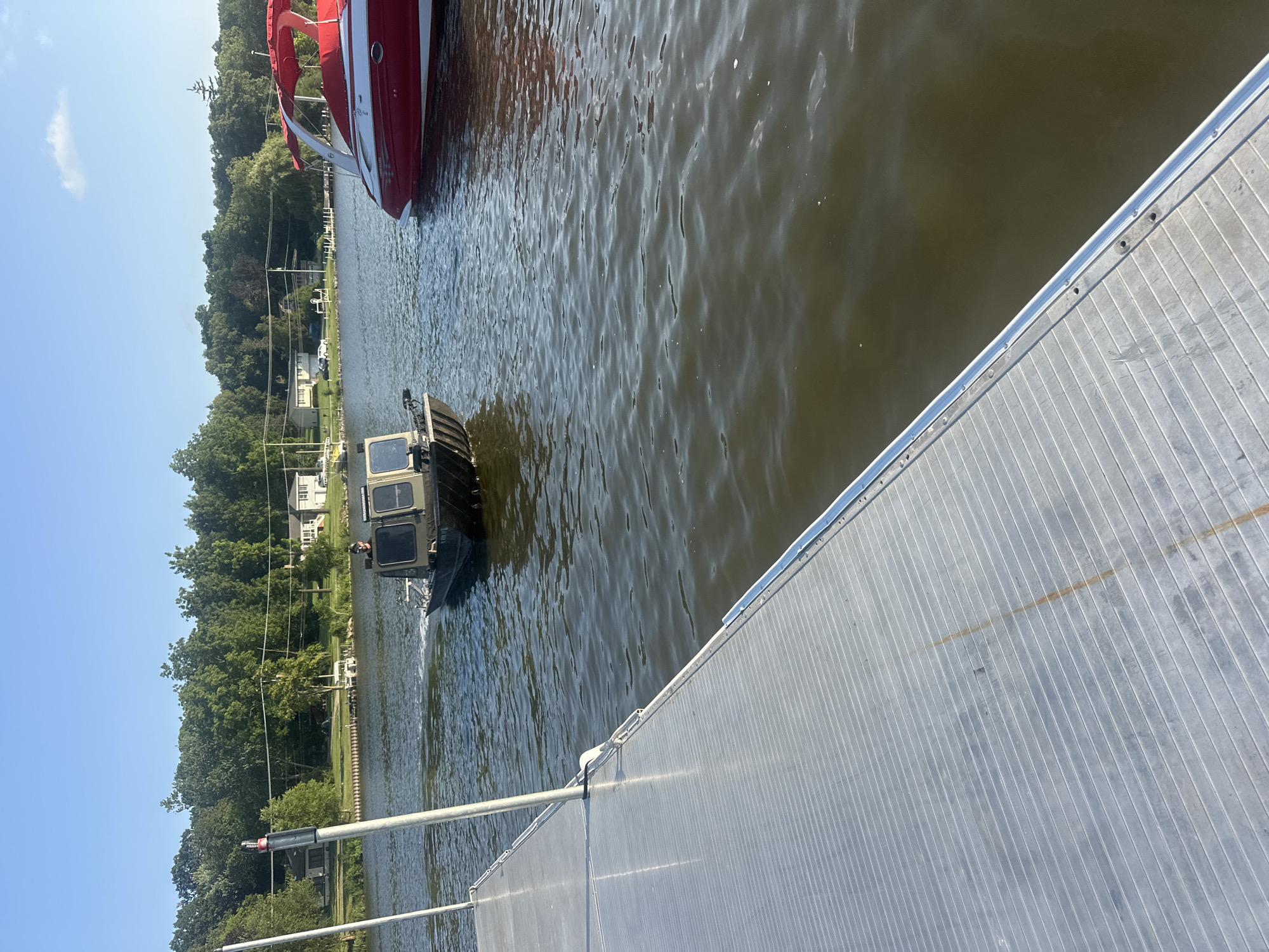 An outdoor, eye-level shot captures a lake with two boats and a dock. On the left, a metal dock juts into the frame. In the center of the lake, a small, dark green boat with a tan cabin and a person inside is moving away from the dock. To the right, a red and white boat with a red canopy is docked. The background features a tree line with houses and a clear blue sky. The scene is set in Algonquin, United States of America.