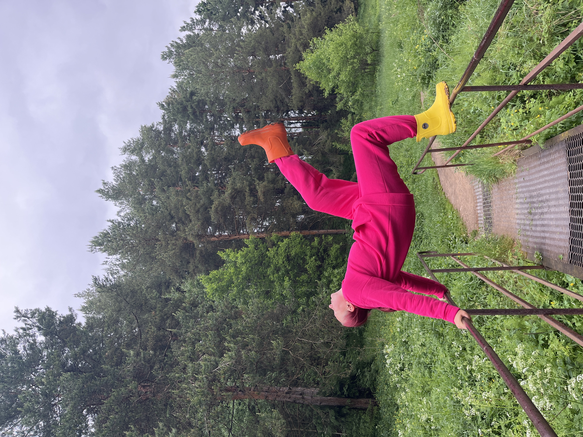 A person is hanging upside down from a metal railing in an outdoor environment. The individual is wearing a bright pink tracksuit, with an orange boot on one foot and a yellow boot on the other. A forest with tall green trees is visible in the background. The railing appears to be part of a bridge or elevated walkway. The location may be in the city of Chaikovsky, Russia.