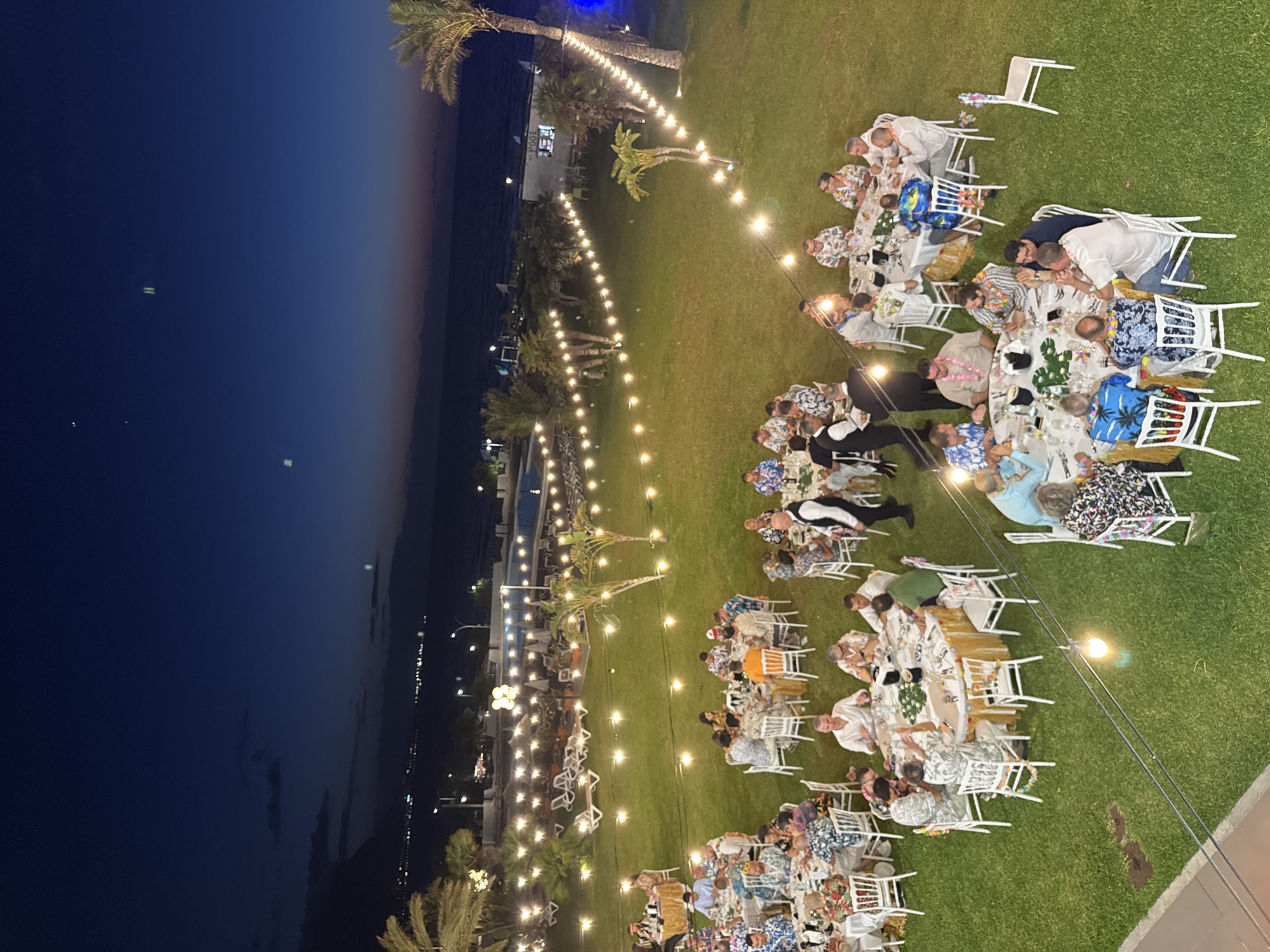 This is a high-angle, wide shot of an outdoor evening event taking place on a manicured lawn near the sea, possibly in Isola delle Femmine, Italy. The scene is illuminated by strings of warm-toned fairy lights draped across the grass and palm trees, creating a festive atmosphere against the twilight sky and the dark expanse of the sea. Several round tables, each adorned with white tablecloths and centerpieces, are set up for dining, surrounded by white chairs. Groups of people are seated at these tables, engaged in conversation and eating. Many of the male guests are wearing brightly colored Hawaiian-style shirts, suggesting a themed celebration. In the background, lounge chairs and umbrellas are visible, along with a swimming pool and buildings, indicating the event is likely at a resort or hotel. The ocean is visible beyond the lawn, with lights from a distant coastline twinkling in the dark. The overall impression is one of a lively social gathering taking place in a scenic coastal location.
