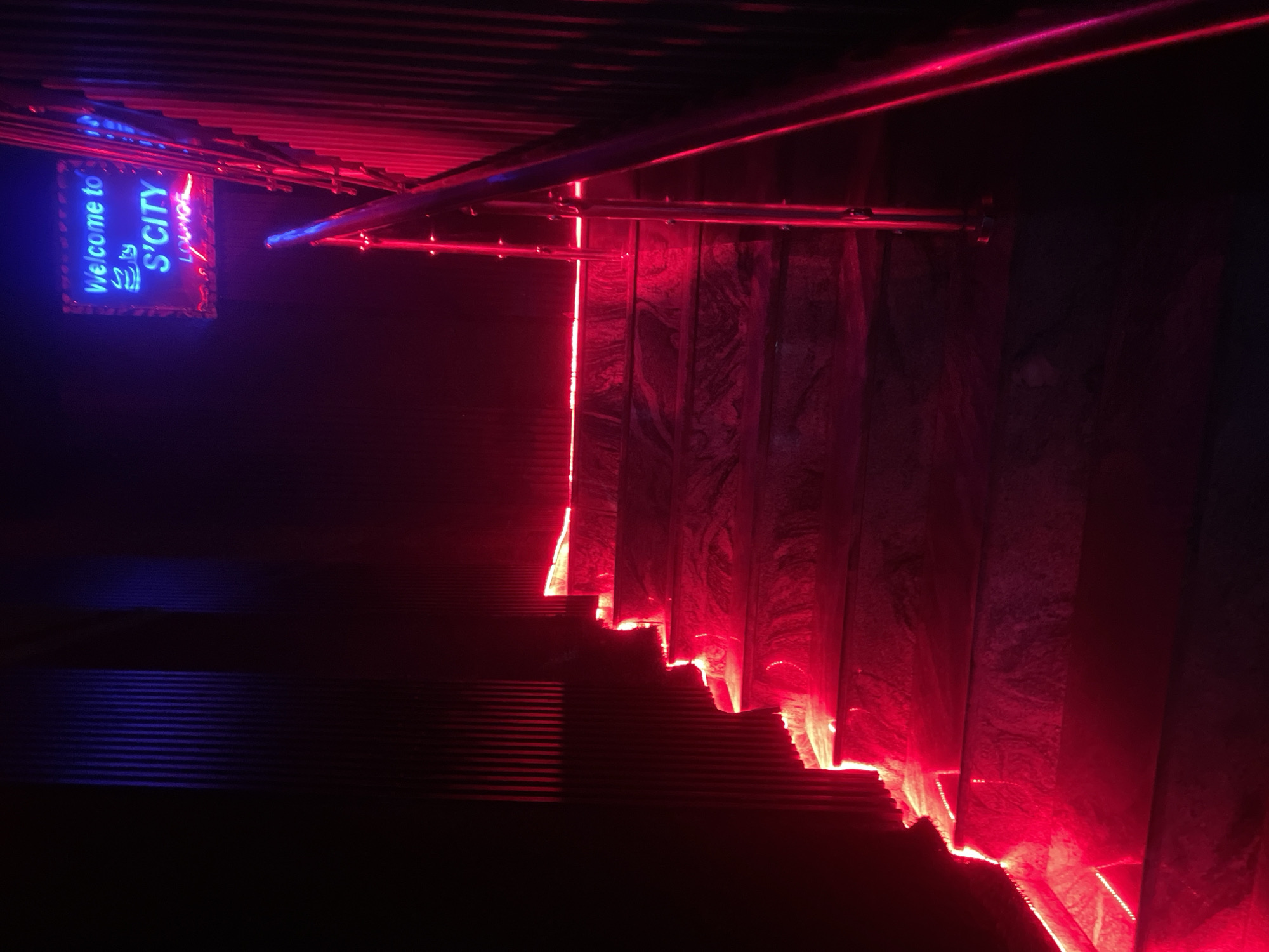 The image depicts a dimly lit staircase with red neon lighting. A blue neon sign in the upper left corner reads "Welcome to S'City Lounge," suggesting a bar or club setting. The staircase is constructed with grey marbled-style material. A stainless steel handrail is visible alongside the stairs. The environment is dark and likely indoors. The location is likely Abuja, Nigeria, based on the sign's reference to "S'City Lounge." There are no people in the image.