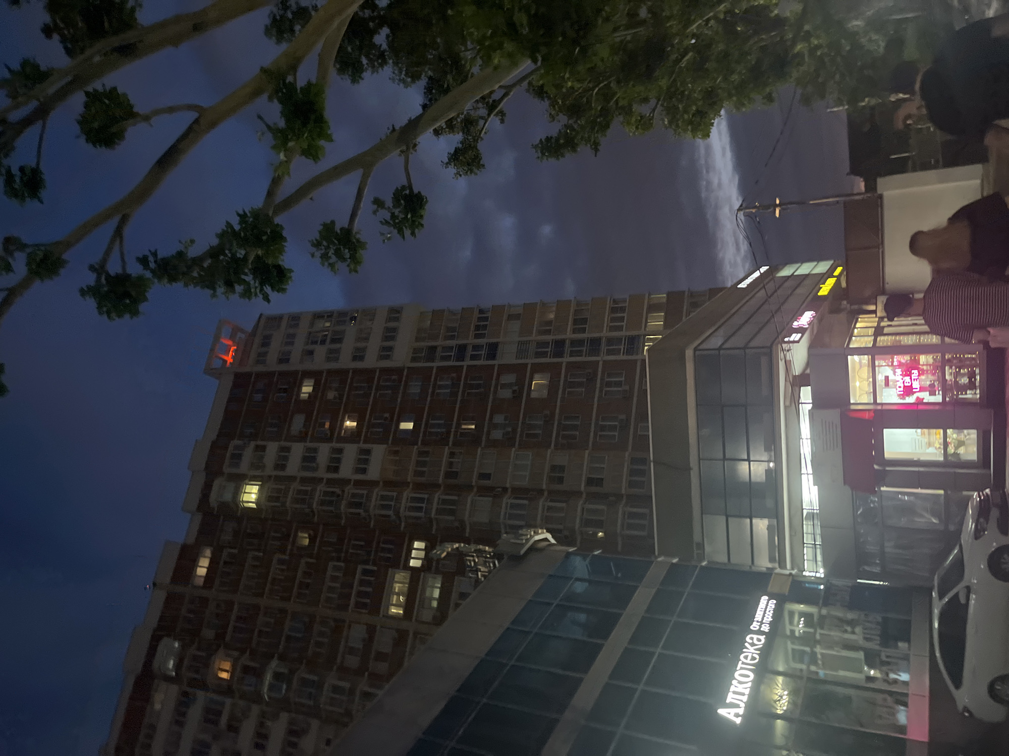 This image depicts a nighttime scene in the city of Krasnodar, Russia. In the foreground, a white sedan is parked on a paved surface, its headlights off. To the left, a building with a glass facade features illuminated signage for "Ал kотeka" (AlkoteKа) and "От винTнoгo до просTого" (From good to simple). Adjacent to this, another section of the building has a red awning over a kiosk displaying flowers and illuminated text. Several people are visible walking away from the camera: a person in a striped shirt, a person with blonde hair, and another person further to the right.
Dominating the background is a tall, multi-story apartment building with numerous lit windows. Above this building, a red illuminated sign with the number "44" is visible. To the right, the branches of a large tree with green leaves are silhouetted against a dark, cloudy sky, suggesting a temperate climate. The overall atmosphere is urban and nocturnal.