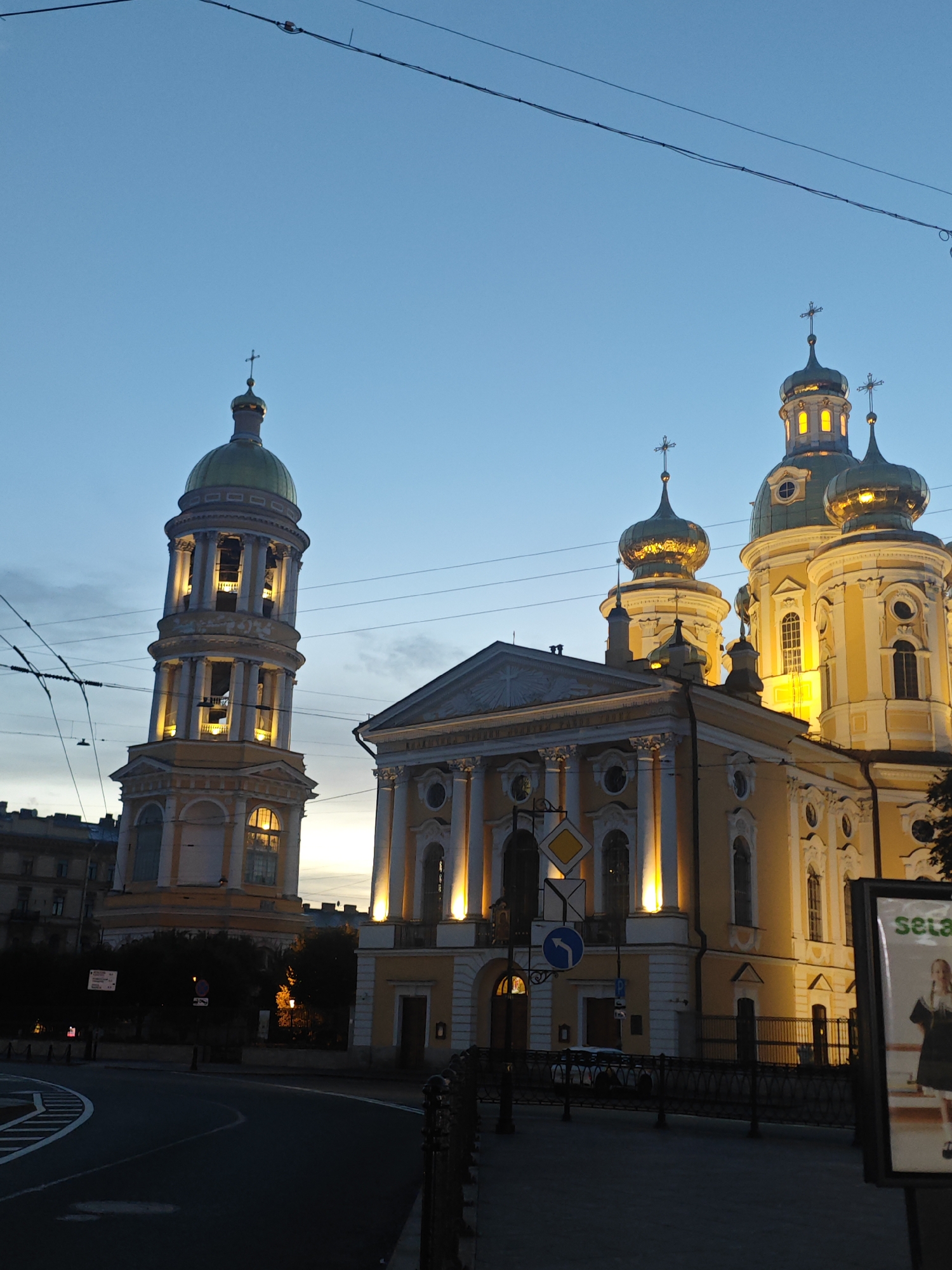 The image shows a street view of a yellow church with golden domes in Saint Petersburg, Russia, at dusk. A tall bell tower is to the left of the church. The sky is a clear blue, with some clouds near the horizon. Electrical wires and tram lines are visible in the sky. A street sign with traffic symbols is in the foreground. There are no people visible in the image. The scene is illuminated by artificial lights from the church and streetlights, creating a warm glow.
