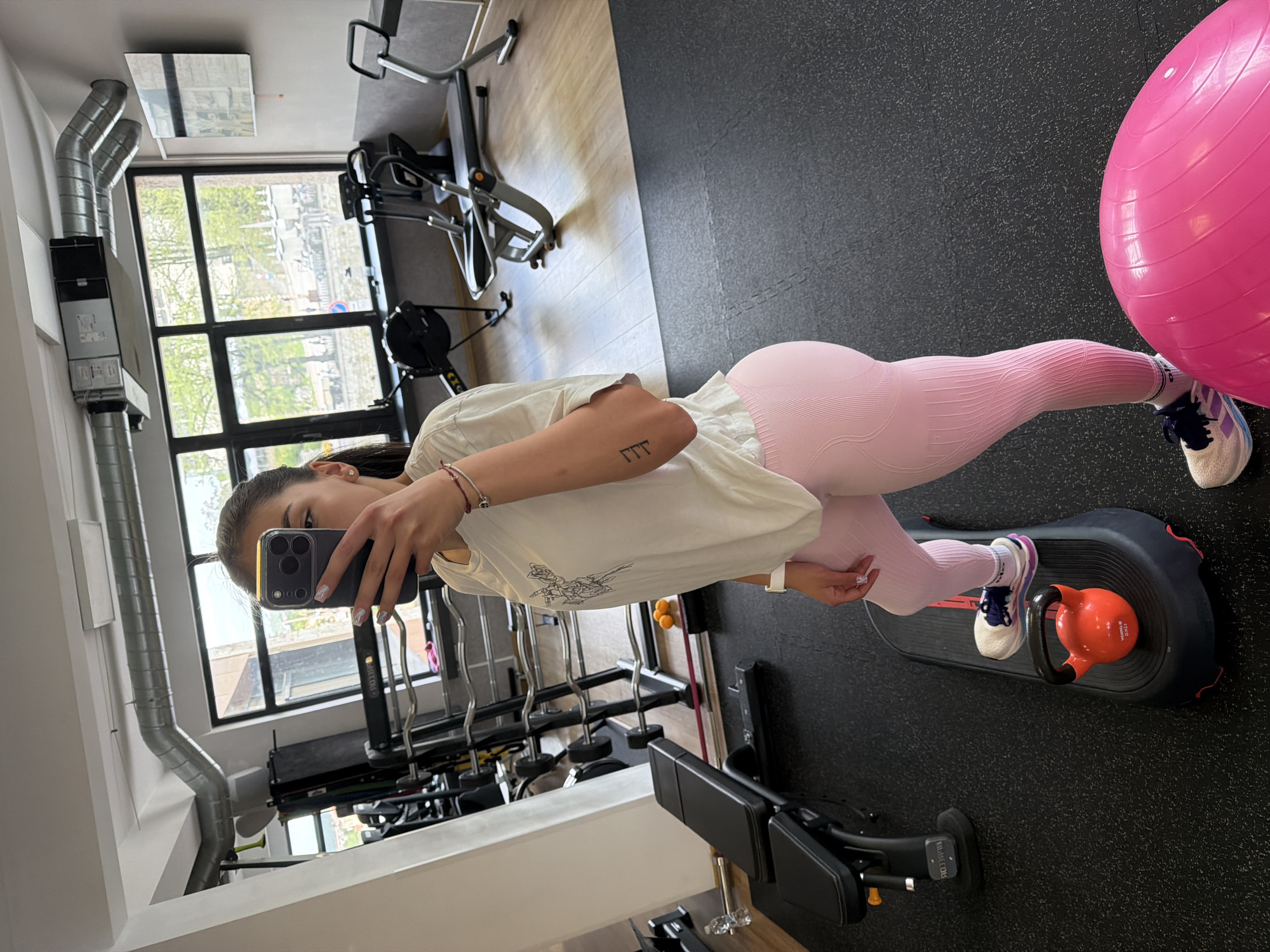 A person is taking a selfie in what appears to be a gym or fitness center. The location is presumably in Gabrovo, Bulgaria.
The individual, a young woman, is wearing pink leggings, a white t-shirt, and athletic shoes. She is holding a smartphone to capture the image.
The environment includes gym equipment such as weight machines, free weights (including a kettlebell), and a large pink exercise ball. There's also a window in the background, which lets in natural light and provides a glimpse of the exterior surroundings. The interior features a combination of wooden and rubber flooring, along with visible ventilation ducts. The image is rotated.