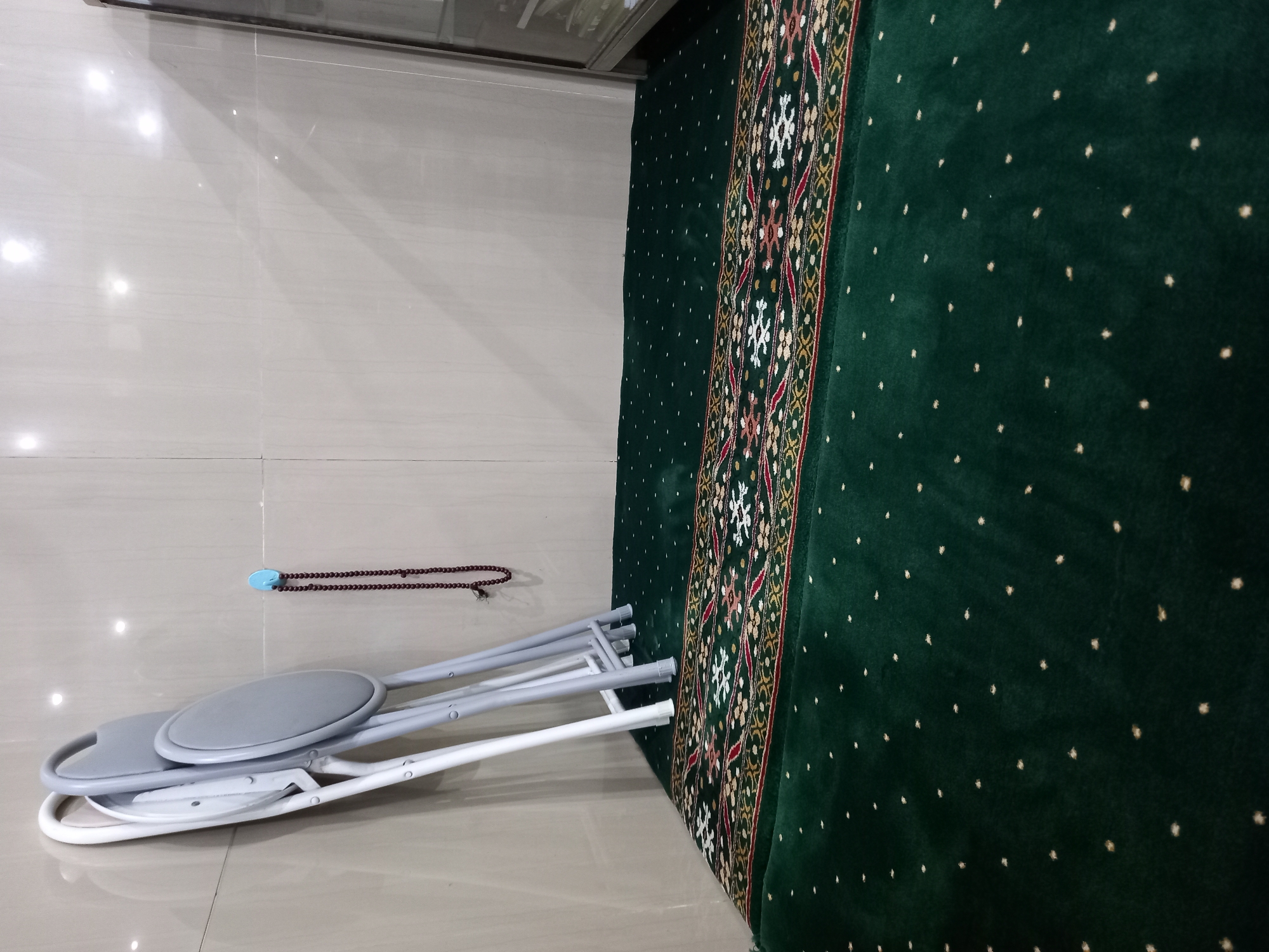 A corner of a room with light-colored, tiled walls and a dark green carpet on the floor. Two folded gray and white chairs are leaning against the wall. A string of dark red prayer beads hangs from a small blue hook on the wall. A decorative, rectangular rug with a red, yellow, and white pattern is placed on top of the green carpet. To the right, a section of a tall, glass-fronted cabinet is visible. The image appears to be taken indoors, likely in a private or communal space in Samarinda, Indonesia, possibly a prayer room.