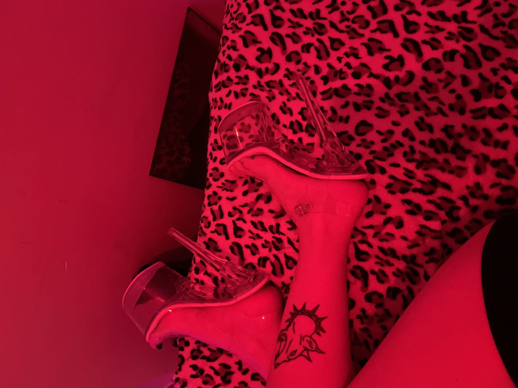 A photograph depicts a scene with a red color cast. A person's leg and foot are visible wearing a clear high-heeled shoe, resting on a leopard print surface. A tattoo is visible on the leg. In the background, there is a red wall. A mirror is also present. The setting is not explicitly identifiable as Buenos Aires, Argentina.