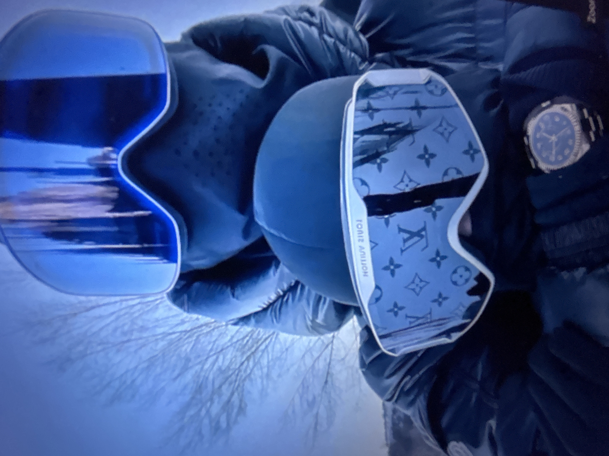 The image displays two individuals, heavily dressed in winter clothing, posing in a close-up outdoor shot. The person in the foreground wears a dark puffer jacket, dark gloves, and a dark balaclava covering their lower face and neck. They are equipped with white-framed ski goggles featuring highly reflective lenses patterned with the Louis Vuitton monogram (interlocking LVs, stars, and quatrefoils), with the brand name "LOUIS VUITTON" legible in reverse on the upper frame. On their left wrist, a silver-toned wristwatch with a dark blue dial and a textured bezel is visible. Behind and slightly above the foreground individual, a second person is similarly dressed in a dark puffer jacket and a balaclava, wearing blue-framed ski goggles with solid reflective blue lenses. A dark cap or beanie is visible between the two figures. The background is blurred, showing a light blue sky and a network of bare tree branches, indicating an outdoor, possibly cold-weather, environment. No specific landmarks, architecture, or unique geographical features are present in the image to identify its location as the city of Burgas, Bulgaria, or any other specific site. The scene is illuminated by cool, ambient light, giving it a predominantly blue cast.