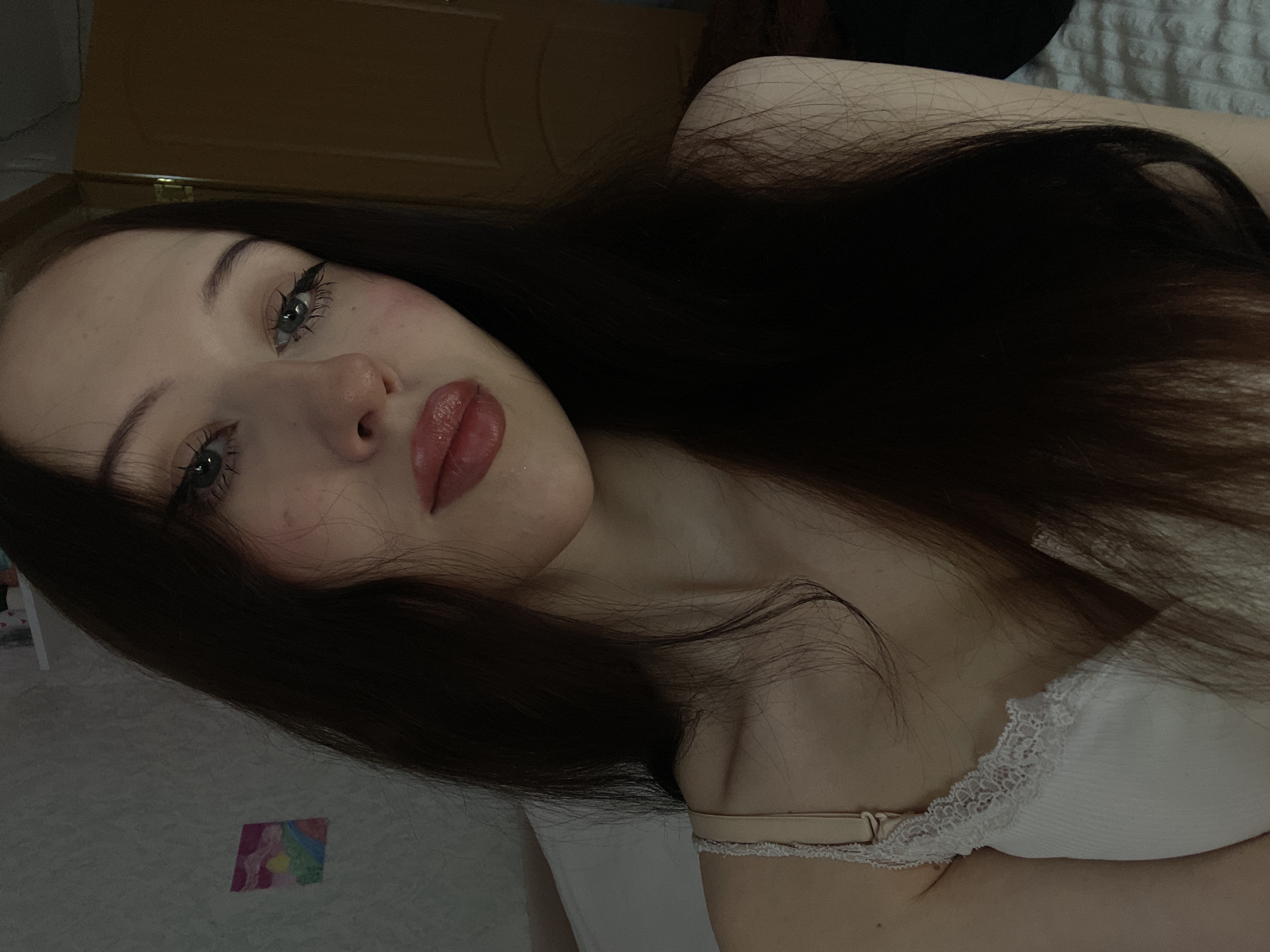A young woman with long, dark hair and striking blue eyes is captured in a close-up portrait. She is wearing a white tank top with lace trim. Her makeup is noticeable, with prominent eyeliner and glossy pink lipstick. The background appears to be an indoor setting, possibly a bedroom, with a plain wall and a door visible. A small, colorful picture hangs on the wall. The lighting is dim, creating a soft and intimate atmosphere. The image does not contain any specific details that would indicate its location in Votkinsk, Russia, or any other city.
