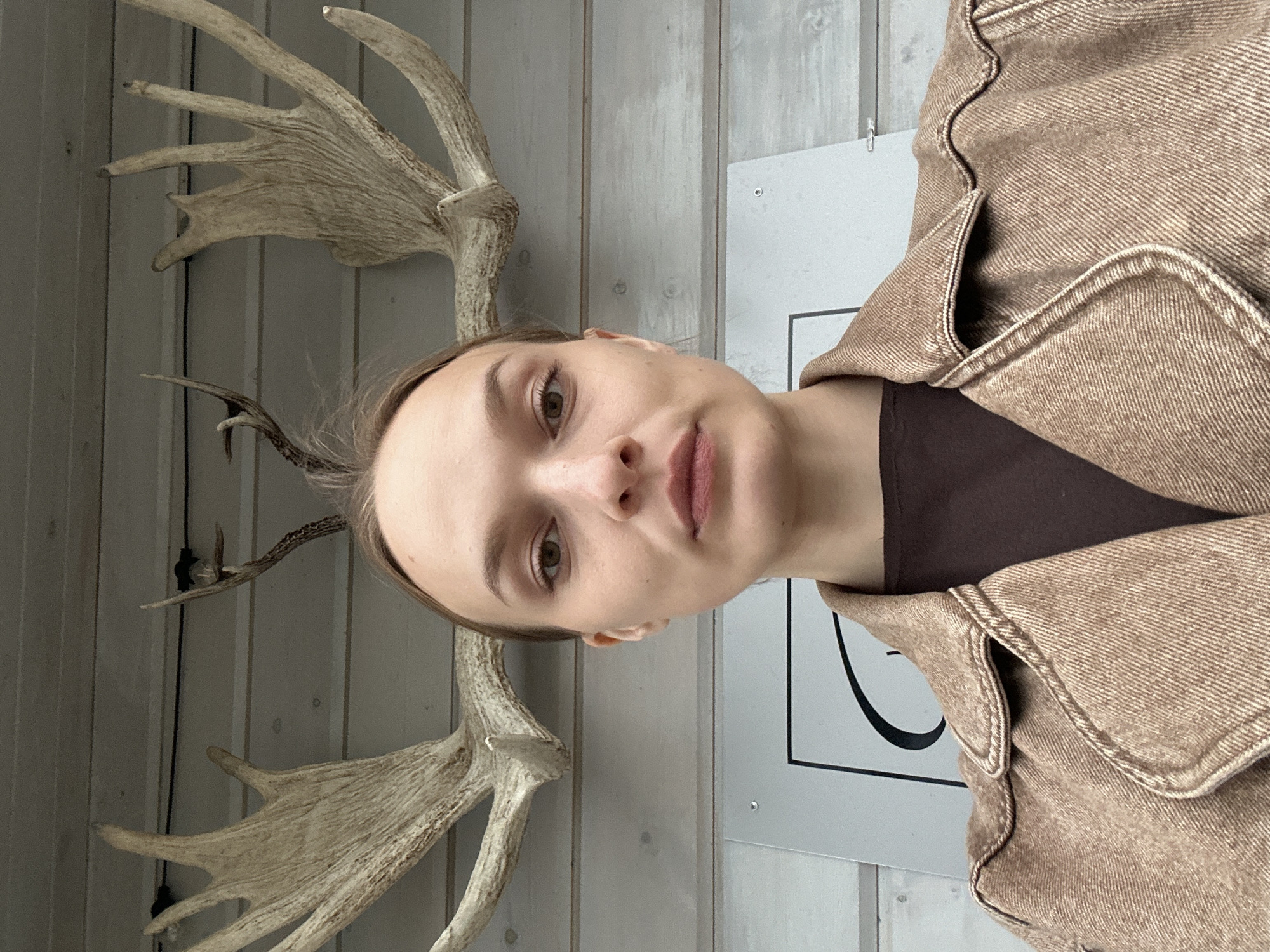 This media file shows a vertical, close-up selfie of a young woman in Karsha, Russia. She is positioned centrally in the frame, looking directly at the camera. The woman has light skin, brown eyes, and light brown hair, with a neutral expression. She is wearing a brown denim jacket over a dark brown top.
Behind her, a wall of light-colored wooden planks serves as the background. Mounted on the wall are large, light-colored antlers, appearing to be from a moose or elk. The antlers are positioned on either side of the woman's head, creating a symmetrical effect. To the right of the woman, a portion of a white framed picture with a black curved design is visible, suggesting an indoor setting.
The lighting appears to be soft and diffused, with no strong shadows, indicating it could be daytime indoors or an overcast day. There are no discernible indicators of the weather or specific time of day beyond that. The overall scene suggests a casual or decorative indoor environment, possibly a home, gallery, or themed establishment. The image is cropped tightly on the woman and the immediate background elements. No text is visible in the image.
