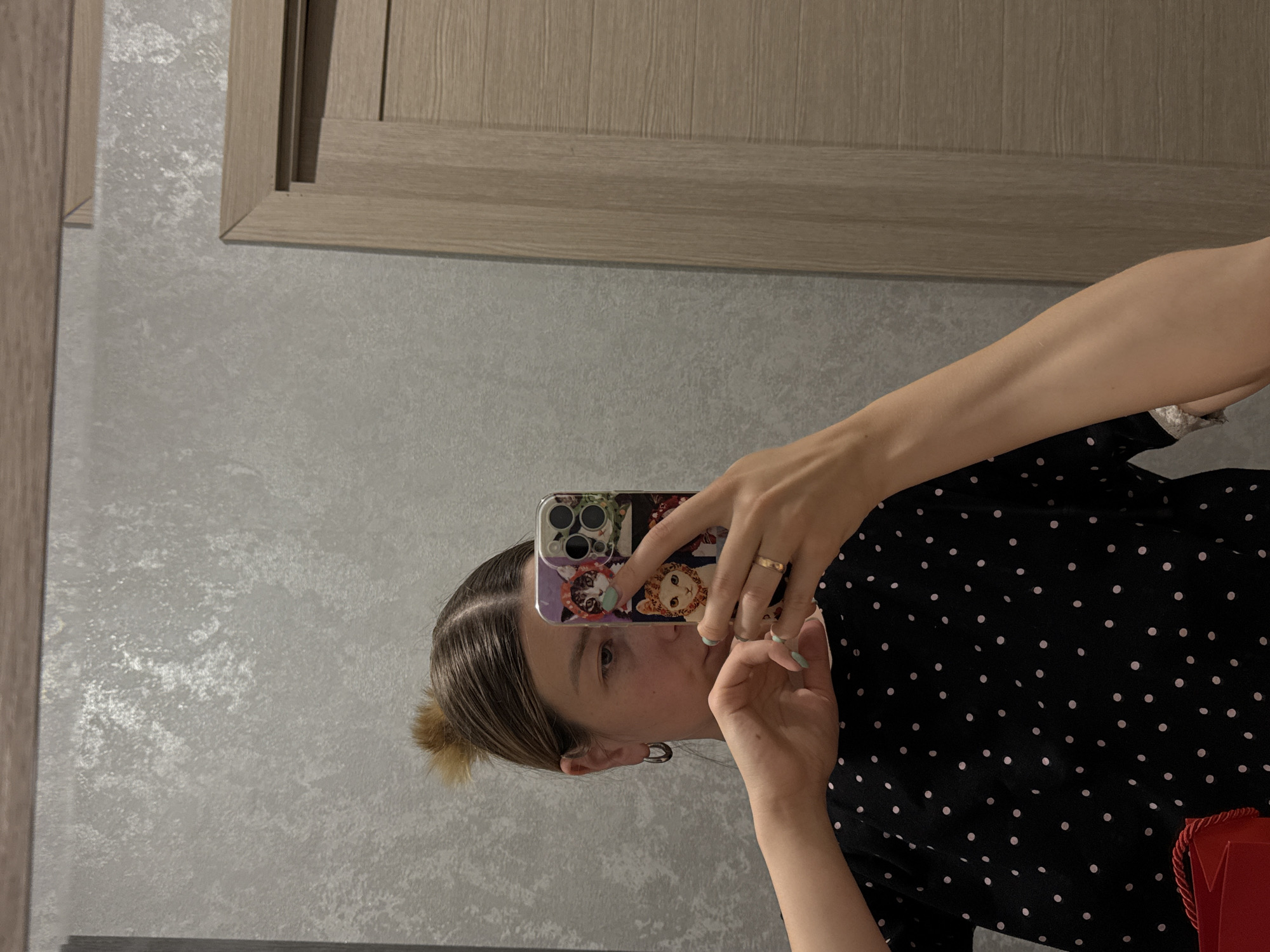 A person is taking a selfie in a mirror. The person is wearing a black and white polka dot shirt. They are holding a phone with a cat-themed case. The background includes a gray wall and a light brown door. The location is indoors. There is no indication the picture was taken in Moscow.