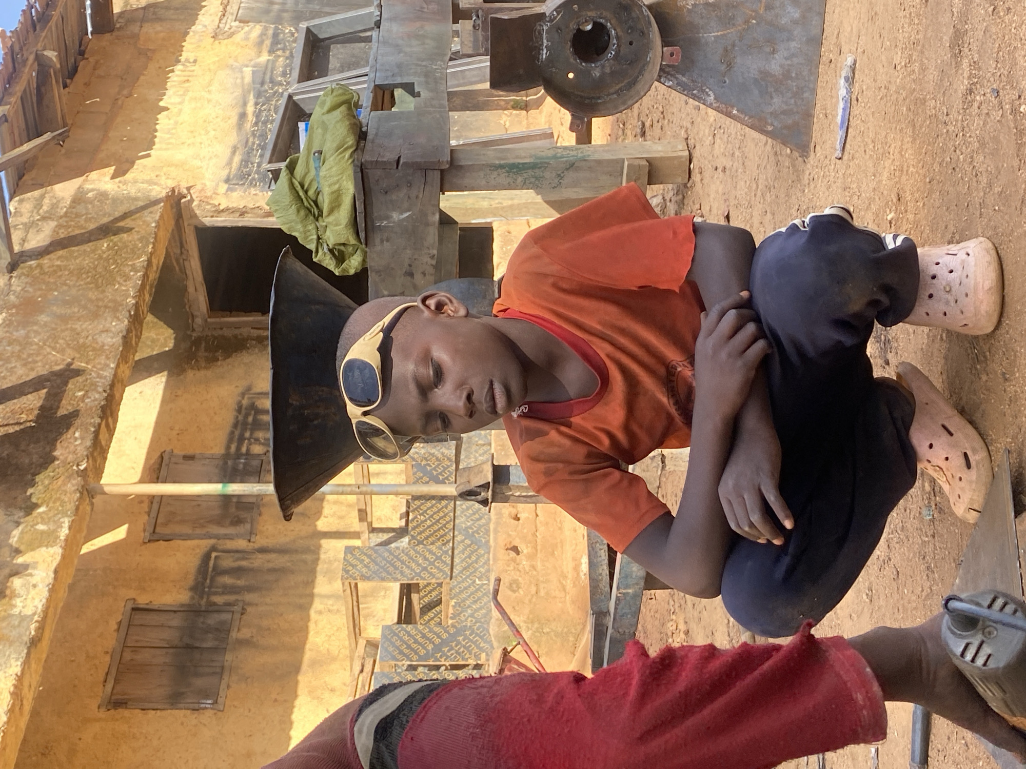 The image shows a young person squatting on the ground in an outdoor setting. The person is wearing an orange t-shirt, dark pants, and light-colored slip-on shoes with holes. Sunglasses are perched on their head. A portion of another person wearing a red shirt is visible in the foreground. The background includes a partially visible building of tan-colored with visible wood framed windows, and construction materials like lumber and plywood, suggesting a construction site. The ground is bare and dusty. Based on the location information provided, this scene is likely in Kuru Dungus, Nigeria.