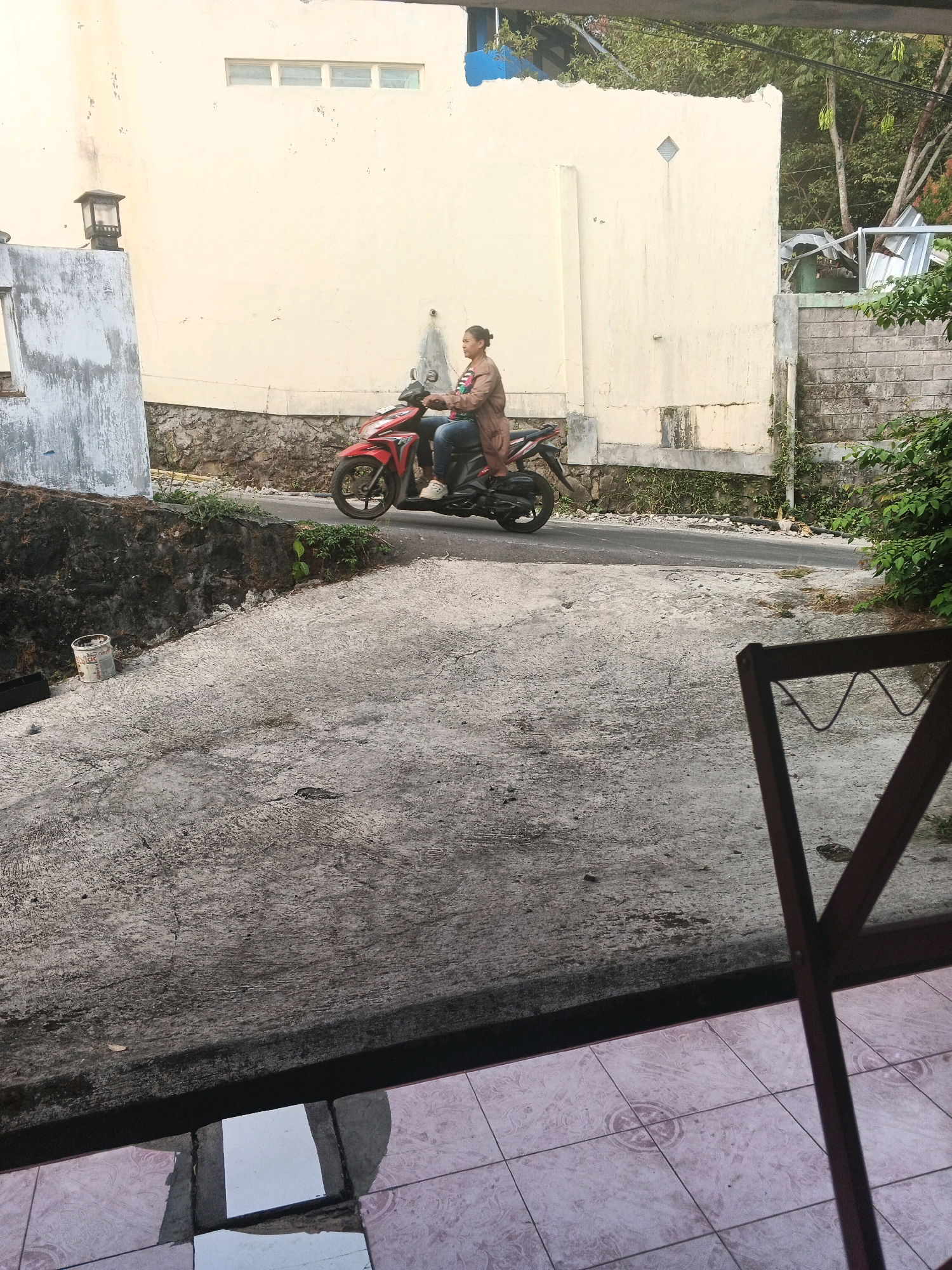 A person is riding a red and black motorcycle on a paved road. The person is wearing a light brown jacket and blue jeans. The motorcycle is in motion, moving from left to right in the frame. The road is bordered by a textured concrete slope on the left and a stone wall with some greenery on the right. Behind the road, there is a pale yellow building with a few rectangular windows. The ground in the foreground is rough concrete, and some patterned tiles are visible at the bottom of the frame, suggesting an outdoor area near a building. The overall scene appears to be an outdoor street view in a residential or commercial area, potentially in Keji, Indonesia, given the context.