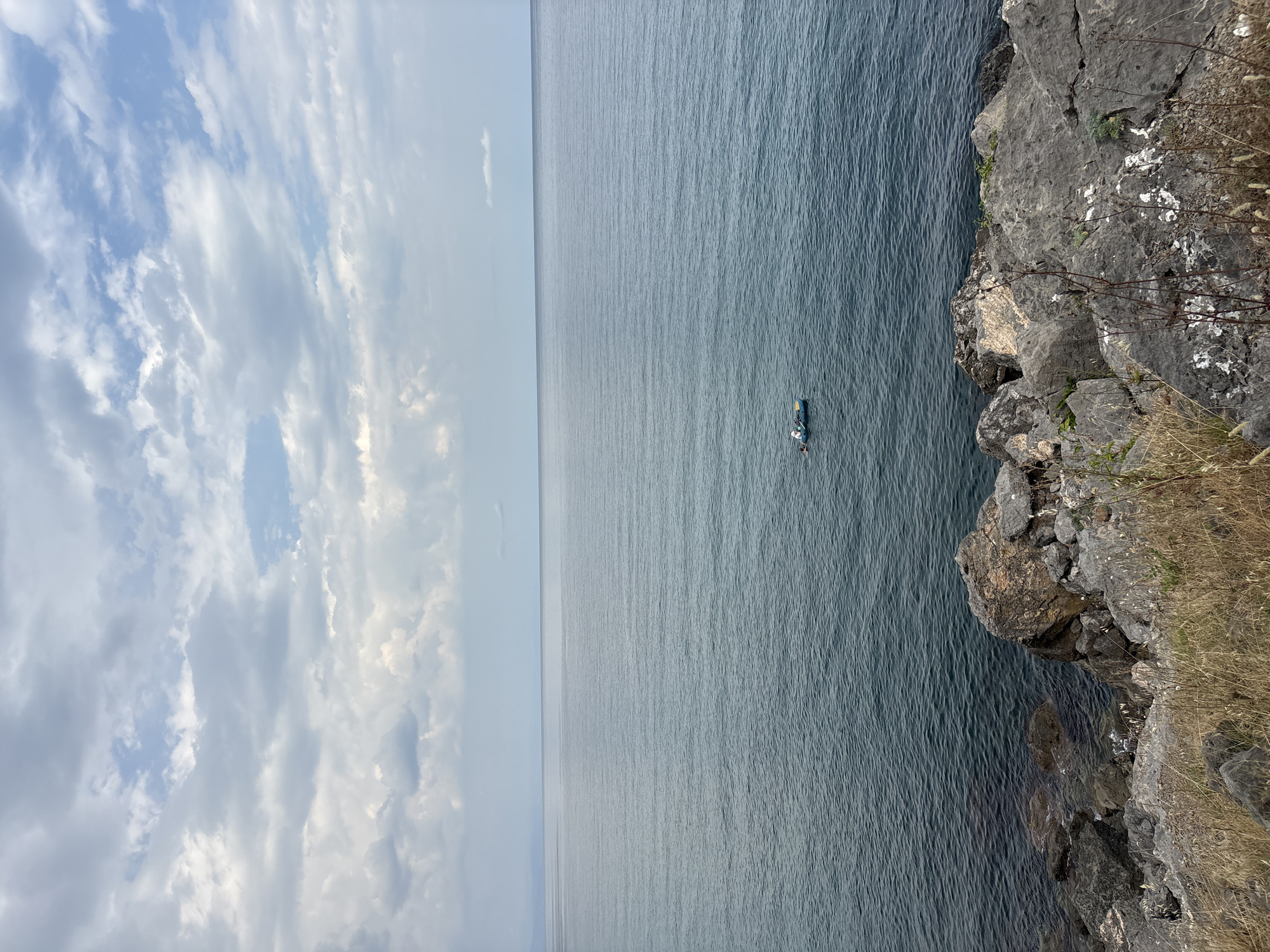 A wide-angle, eye-level, outdoor, full shot captures a calm sea under a cloudy sky, with a rocky shoreline in the foreground. The sea is a deep blue, with subtle ripples indicating minimal wave activity. In the distance, a small, light blue boat with a white-capped person is visible on the water. The sky is filled with a mix of light and dark grey clouds, with patches of pale blue sky peeking through. The foreground is dominated by large, grey and brown rocks, with dry, light brown grass and some sparse green vegetation growing between them. The scene appears tranquil and serene, with the vastness of the sea and sky emphasizing a sense of solitude. The location is not identifiable as Bondarenkovo, Ukraine.