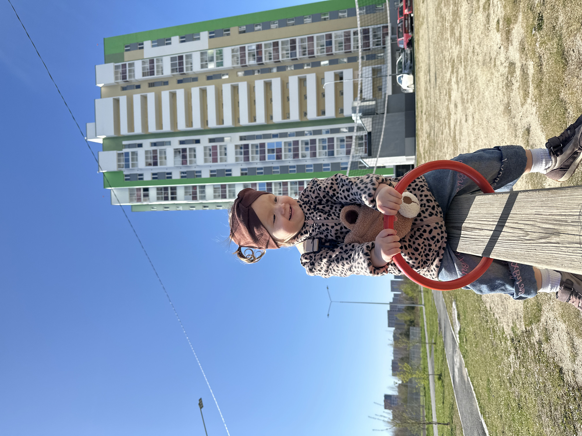 The image shows a young girl on a playground seesaw in Yekaterinburg, Russia. She is wearing a brown hat, a leopard-print jacket, blue jeans with writing on them, white socks, and brown sneakers. She is holding a brown teddy bear. Behind her, there is a tall building with green, beige, and white sections, suggesting it's a modern apartment complex. A chain-link fence and a white vehicle are visible behind the girl and the building. The ground is a mixture of sand and grass. The sky is blue with a faint contrail from a jet. The lower part of the image shows a walkway, grass and some trees, and buildings in the distance.