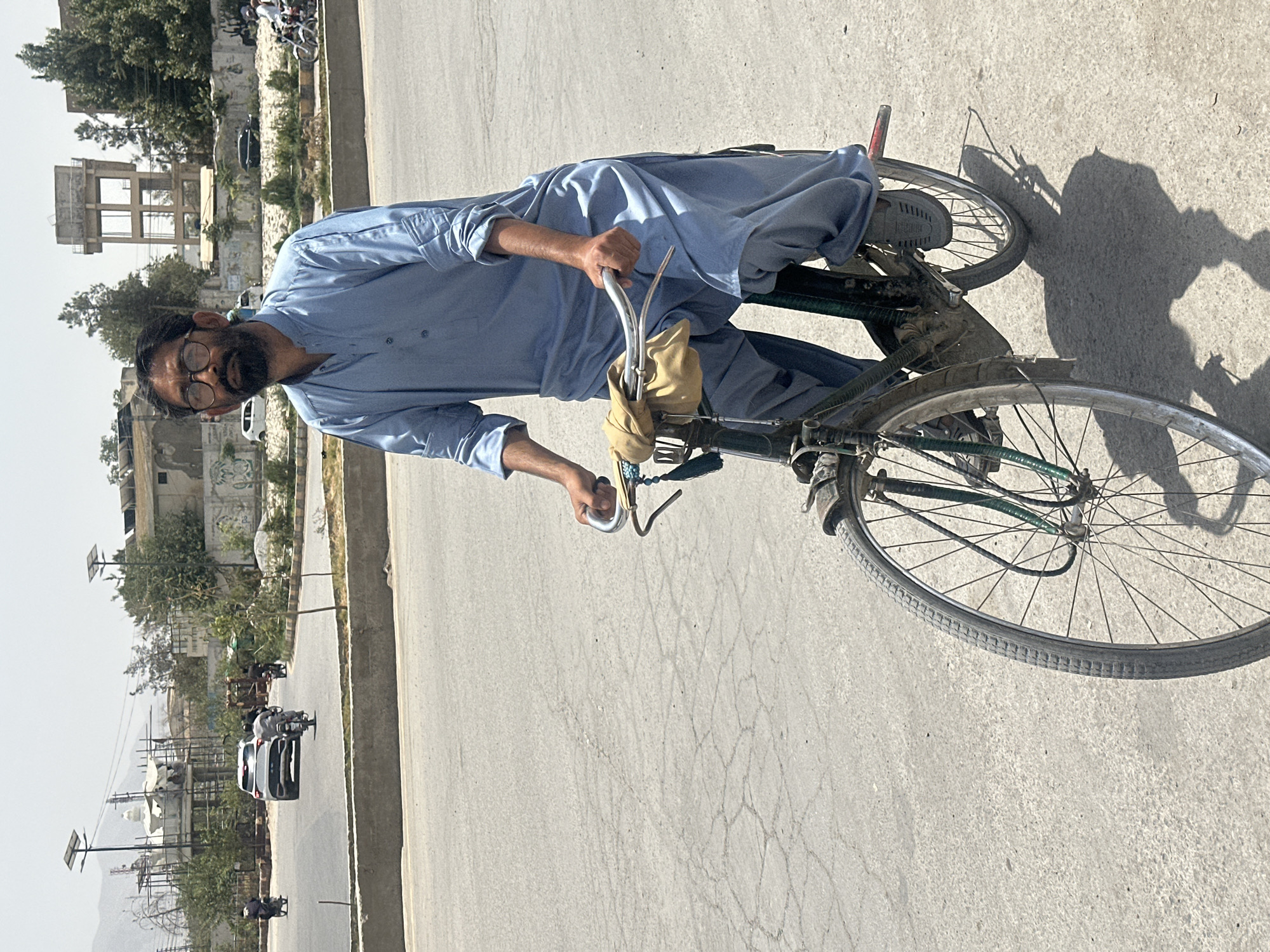 The image depicts a man in a light blue shalwar kameez riding a bicycle on a paved road in Quetta City Tehsil, Pakistan. The man has dark hair, a beard, and is wearing glasses. He is looking forward, and his hands are on the handlebars. A light brown cloth is tied to the handlebars, and a string of blue beads hangs from it. The bicycle is old and appears to be in use. In the background, there are buildings, trees, and a water tower. Several other vehicles, including motorcycles and a white SUV, are visible on the road. The sky is clear and the sun is shining, casting shadows on the ground. The road is cracked in places, indicating it is an older road surface. The overall environment suggests a warm, sunny day in a city.