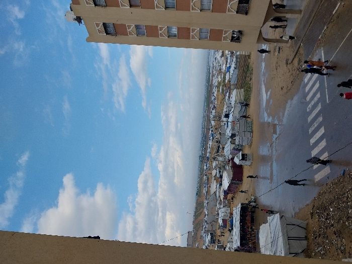 The image displays an elevated view of a section of Khan Yunis, Palestinian Territory, featuring a street, a permanent building, and a vast displacement encampment. The sky above is partly cloudy, with significant patches of blue visible alongside scattered white cumulus clouds.
In the immediate left foreground, a section of a light-colored building, from which the photograph appears to be taken, is visible. To the right, a five-story tan-colored building with distinctive white decorative arch-like frames around its windows stands, featuring a satellite dish on its roof and an open, covered pedestrian area on its ground floor.
A wet, dark asphalt street with a marked white pedestrian crosswalk (zebra crossing) runs horizontally across the mid-ground. Numerous puddles are visible on the street surface and the adjacent sandy, muddy ground, indicating recent precipitation. Beyond the street, stretching into the background, is an extensive, densely populated encampment composed of makeshift shelters. These shelters are constructed from various materials, including white tarpaulins, corrugated metal sheets, and other temporary materials, packed closely together and extending towards a hazy horizon.
Approximately two dozen individuals are present. Several pedestrians, including adults and children, are observed walking across the wet street, with some utilizing the zebra crossing. Others are visible walking on the sandy, muddy terrain adjacent to the encampment or standing beneath the covered ground floor of the building on the right. On the left side of the street, bordering the encampment, a market stall or vendor setup under a white canopy is discernible. A dark utility cable or line diagonally traverses the frame from the upper left.