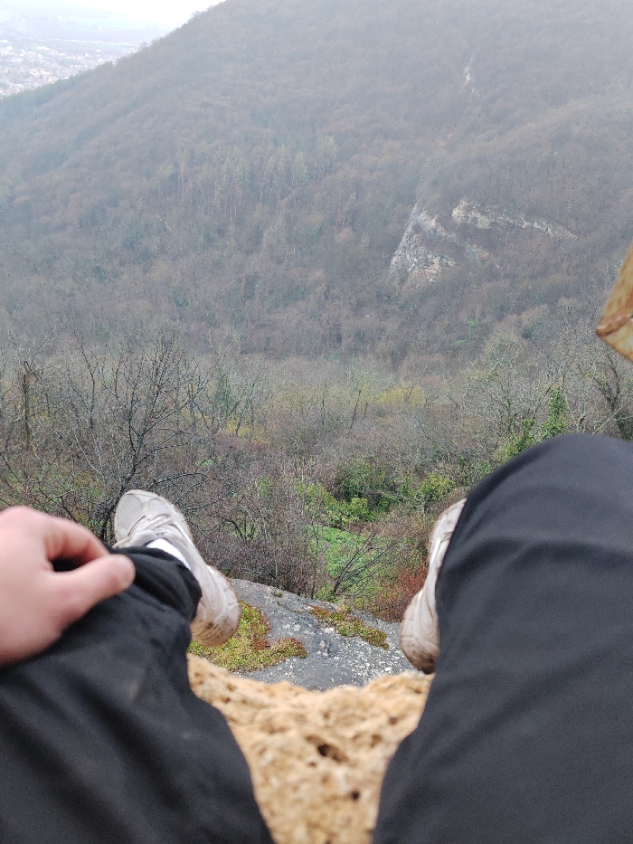 The image presents a first-person perspective, looking downward from a high vantage point over a forested valley. In the immediate foreground, a person's legs and left hand are visible, indicating the individual is seated or lying at the edge of a rock formation. The person wears black trousers and light-colored athletic shoes, stained with dirt. The ground beneath the person is coarse, light brown, porous rock, with patches of darker rock and green moss visible directly below the feet.
The view extends over a steep, densely wooded valley, characterized by predominantly bare deciduous trees, suggesting late autumn or winter. Patches of brighter green vegetation are visible in the valley floor. On the opposing hillside, evergreen trees are interspersed among the deciduous ones, and a prominent, lighter-colored rock face or cliff formation is visible on the right side of the slope. The distant background, visible in the upper left, shows a hazy outline of a cityscape, consistent with Shumen, Bulgaria, as viewed from the Shumen Plateau. The sky is overcast and hazy, diffusing the light and contributing to a muted color palette. A small, rusted metal object is partially visible in the upper right corner of the frame.