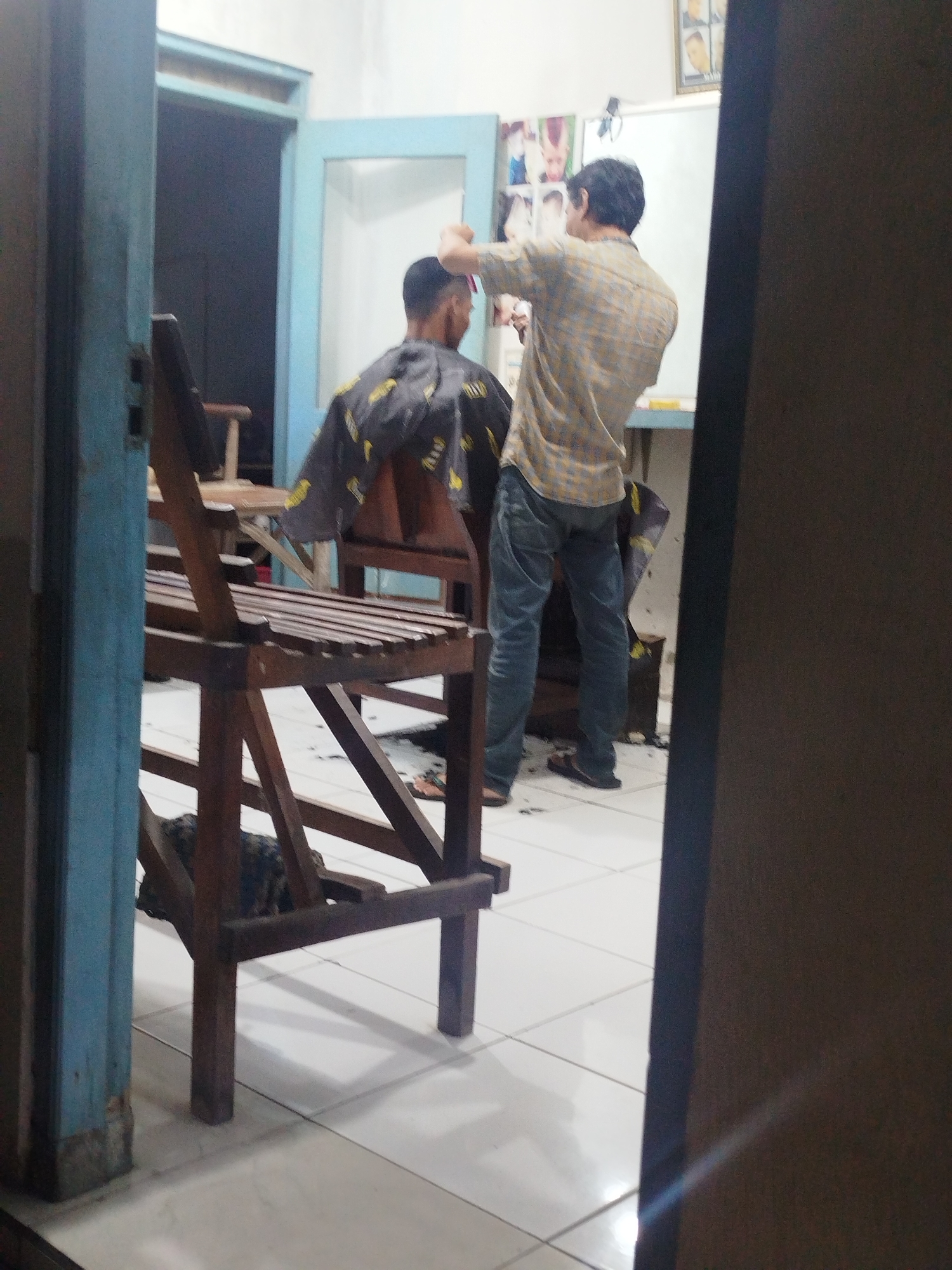The image depicts a barbershop in Tonjung, Indonesia. A man is getting a haircut from another man. The barber is wearing a plaid shirt and jeans, while the person receiving the haircut is covered with a cape. The barbershop appears to be a simple establishment with wooden furniture and tiled floors. The lighting is dim, suggesting it might be late in the day or the shop does not have strong artificial lighting. There are posters on the wall behind the barber, possibly of hairstyles. The overall impression is of a functional, local barbershop.