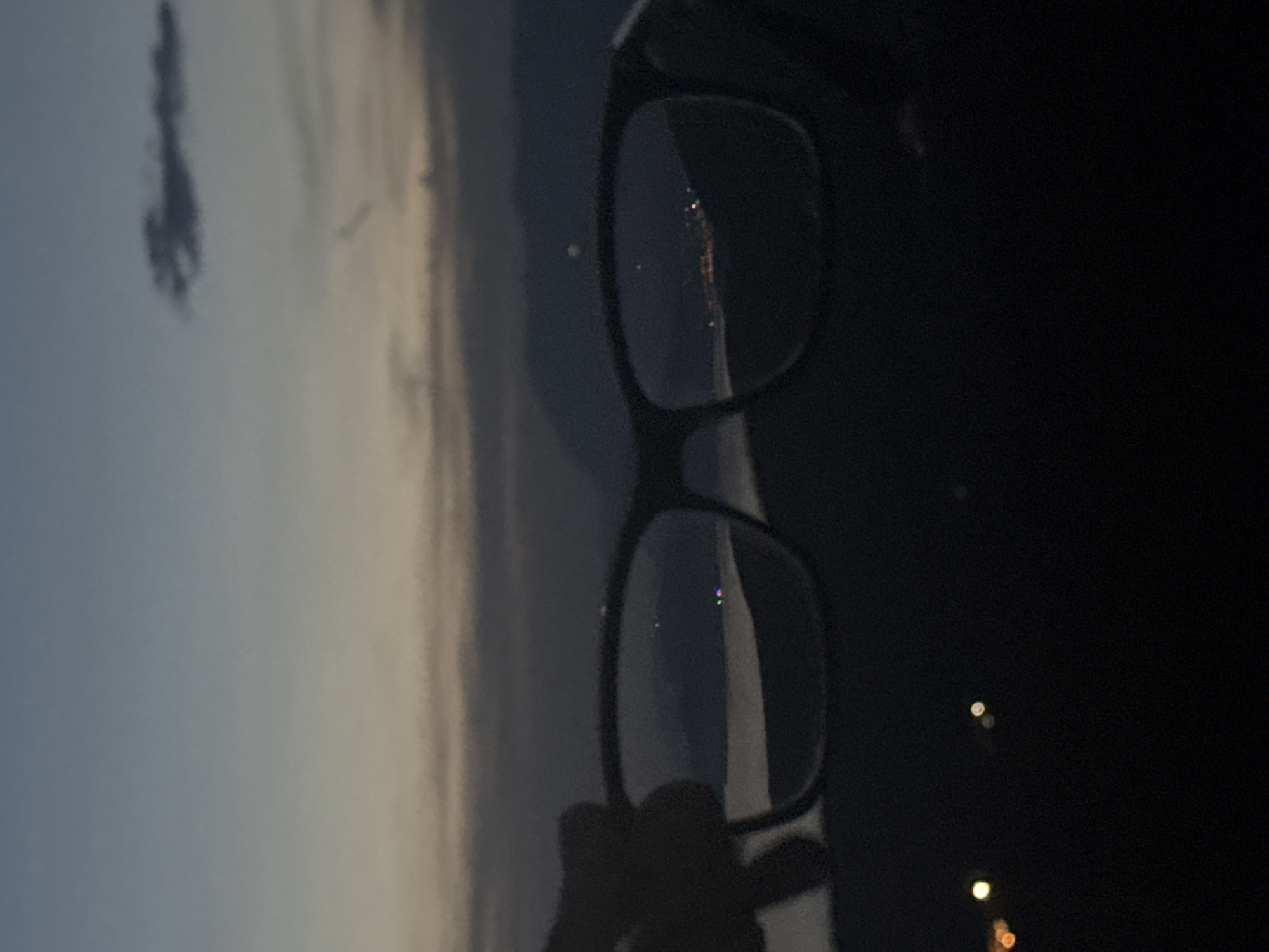 The image shows a pair of black-rimmed eyeglasses held against a landscape featuring a blue sky, clouds, and a faint shoreline. The eyeglasses' lenses reflect fragmented views of the scene, including the distant shoreline. In the lower right, the background is dark, with blurred lights. The image appears to be taken at twilight or dawn, with the sky transitioning from light to dark. The specific location within Kovačko Polje, Montenegro, cannot be determined from the image. There are no people clearly visible. The action is holding the eyeglasses. The interaction is the eyeglasses positioned against the background landscape. The environment is a landscape with a coastline and the time of day appears to be twilight or dawn.