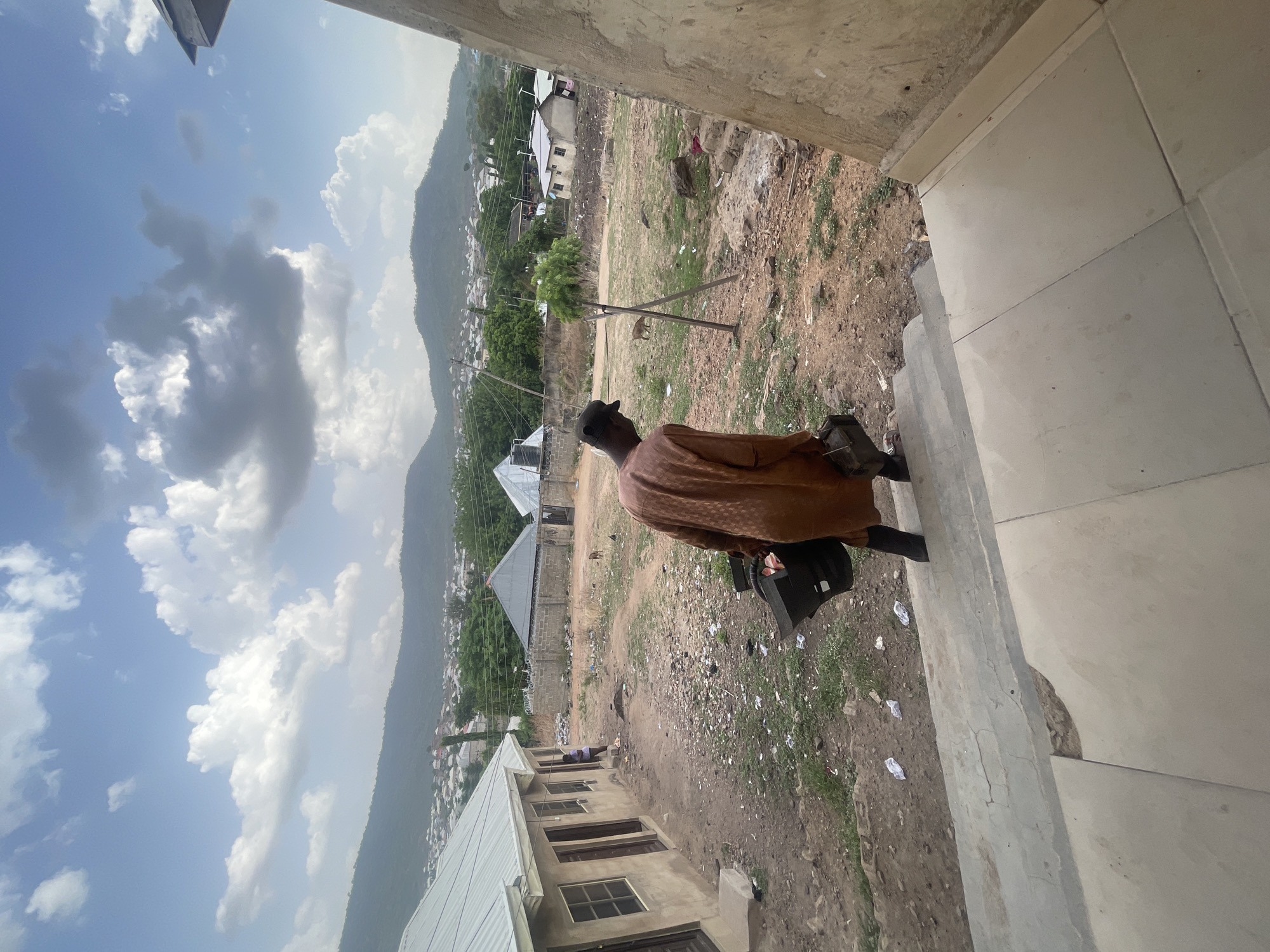 This media file captures an outdoor scene in Lokoja, Nigeria, from an elevated perspective, likely a balcony or building rooftop. The image is oriented vertically (originally rotated 90 degrees clockwise).
In the foreground, a person is seen from behind, facing away from the camera and looking out over the landscape. They are wearing a dark cap and a brown, somewhat textured garment, possibly a traditional top, and are carrying a black bag or briefcase in their left hand. The person stands on a concrete tiled surface, bordered by a concrete ledge or wall on the right.
Beyond the person and the immediate concrete structure, the scene opens up to a view of a developing residential area. The ground is largely unpaved and dusty, with patches of sparse green vegetation and scattered debris. Several houses with corrugated metal roofs are visible, some appearing unfinished or in various stages of construction. Utility poles and power lines crisscross the middle ground.
In the distance, a range of prominent hills or mountains defines the horizon, partially obscuring more residential buildings nestled at their base. The sky above is bright and clear with scattered white and grey cumulus clouds, indicating a sunny daytime. The overall impression is that of a semi-urban or rural environment undergoing development, set against a natural, mountainous backdrop. No visible text is present in the image.