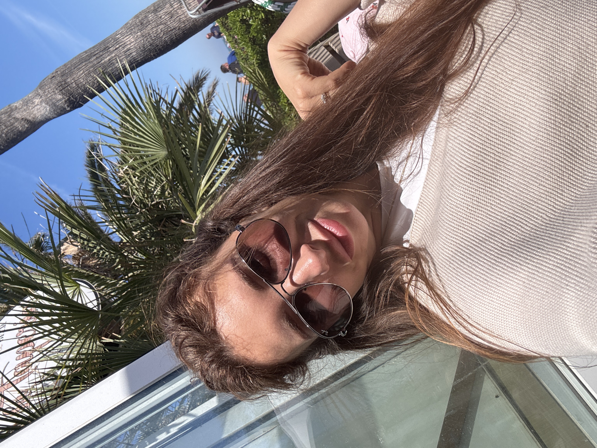 A selfie taken outdoors in Valencia, Spain, presents a woman wearing sunglasses. The backdrop reveals palm trees and blue sky.