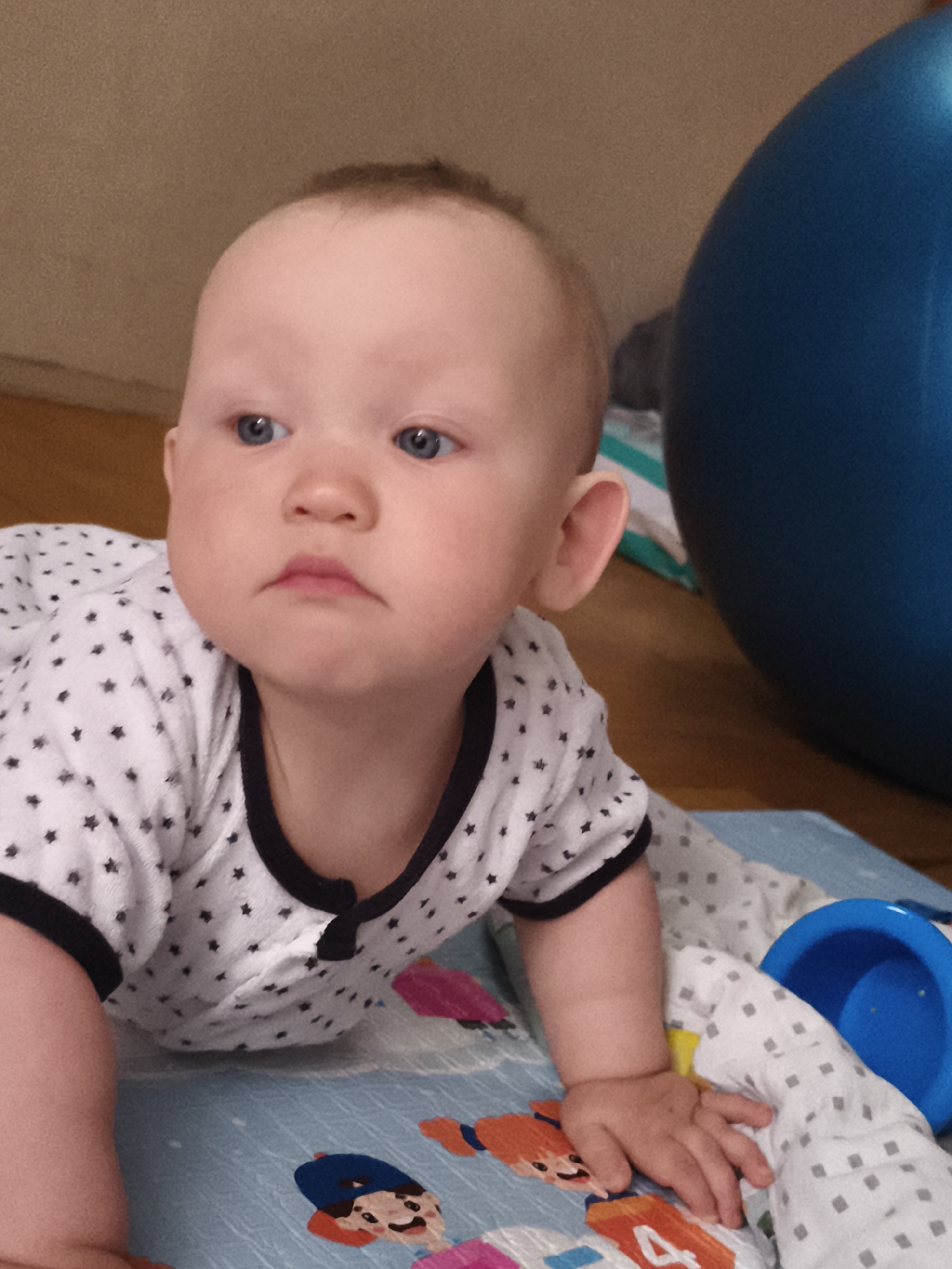 A baby is crawling on a blue and white patterned mat. The baby is wearing a white outfit with small black stars and a black trim around the neck. A large blue exercise ball is visible in the background. The scene takes place indoors on a wooden floor. The specific location in Chaikovsky, Russia cannot be determined from the image alone.