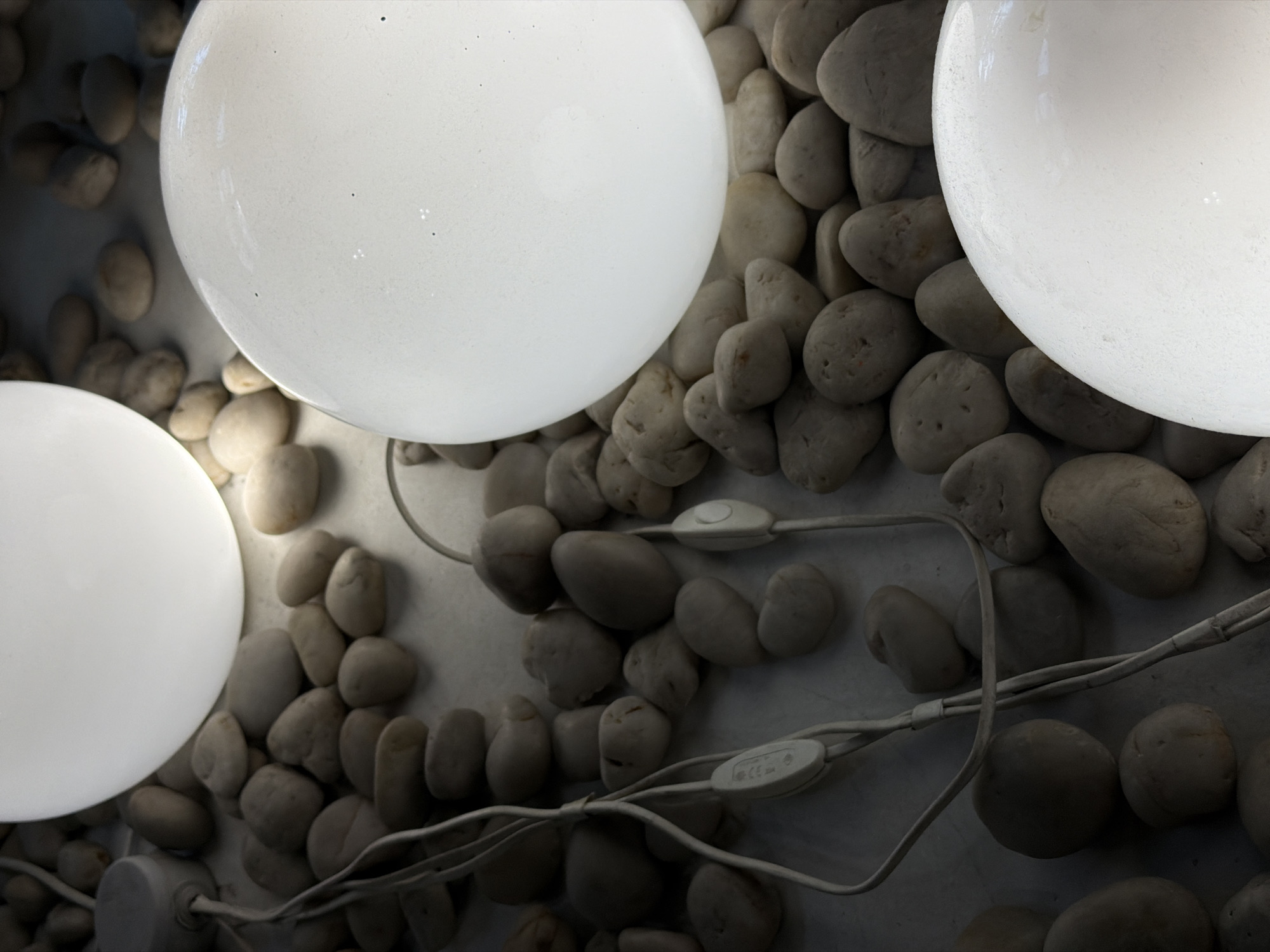 A close-up, indoor shot shows three illuminated, frosted glass sphere light fixtures resting atop a surface covered with smooth, light gray river rocks. Electrical cords, also gray, snake among the rocks, featuring white control switches. The scene is generally dark, with the white spheres providing the primary light source.