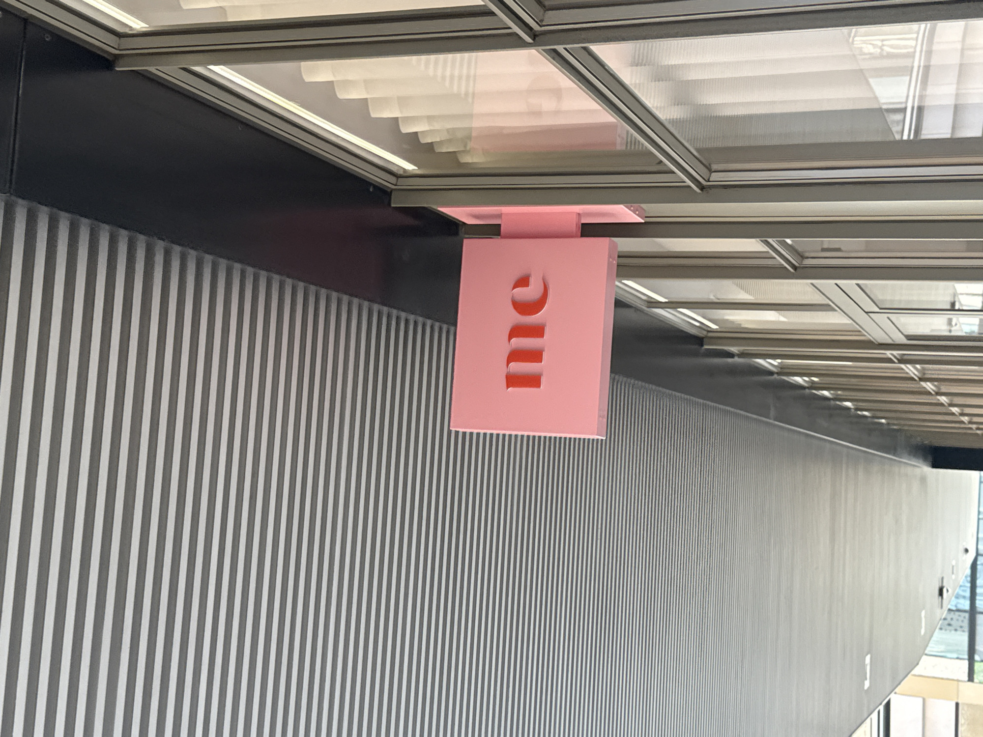 The image depicts a pink cuboid sign with the word "me" cut out in a stylized font, hanging from the ceiling within what appears to be an interior space. The background consists of a wall with vertical gray and white stripes. The ceiling is a grid of clear, corrugated panels set in a metal framework. The location could be in London, United Kingdom, but there are no definitive details to confirm this.