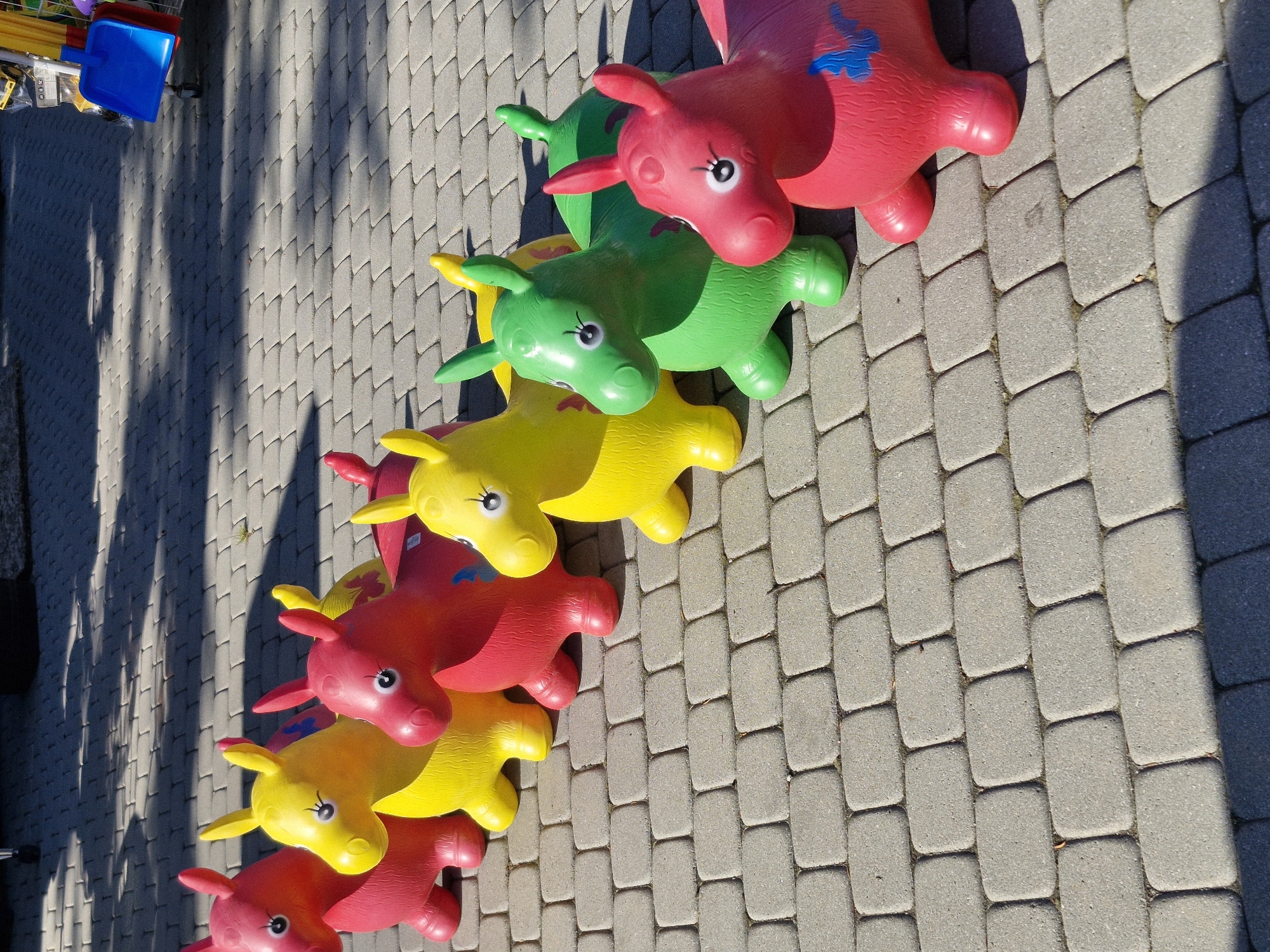 An outdoor scene features seven inflatable horse-shaped bouncing toys arranged diagonally in a line on a paved surface, extending from the lower left foreground to the upper right background. From left to right, the horses are colored red, yellow, red, yellow, green, and two more red horses, the last of which has a blue butterfly decal on its side. All horses share similar features, including distinct black-and-white eyes and blush marks on their cheeks. The ground consists of grey rectangular interlocking pavers. Strong sunlight casts elongated shadows from the horses onto the pavement, suggesting either morning or late afternoon. In the upper right background, a blue plastic scoop and other yellow items are partially visible, suggesting a display or stall. The location is an unspecified outdoor paved area in Unknown, Unknown.