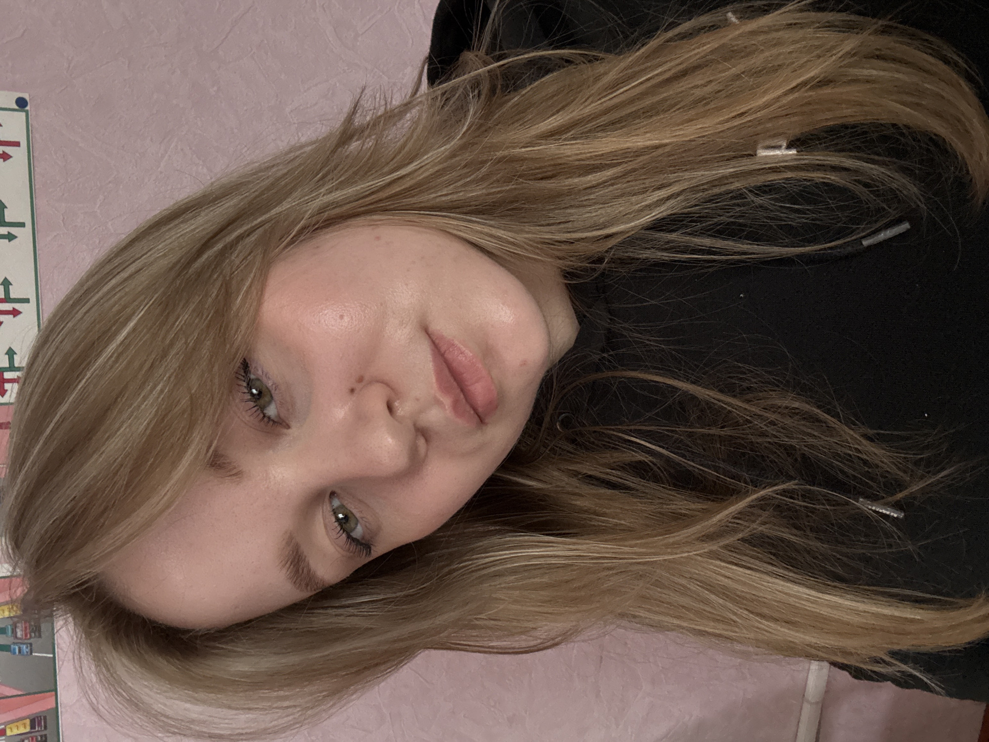 A young woman with blonde hair and green eyes poses for a photo. She is wearing a black hoodie. The background is a pale pink wall with a section of a colorful picture frame or poster visible in the upper left corner. The location is likely inside a building, potentially a private residence, but the city of Chaikovsky, Russia cannot be independently verified from this image alone. No actions or interactions can be observed beyond the subject posing for the photo.