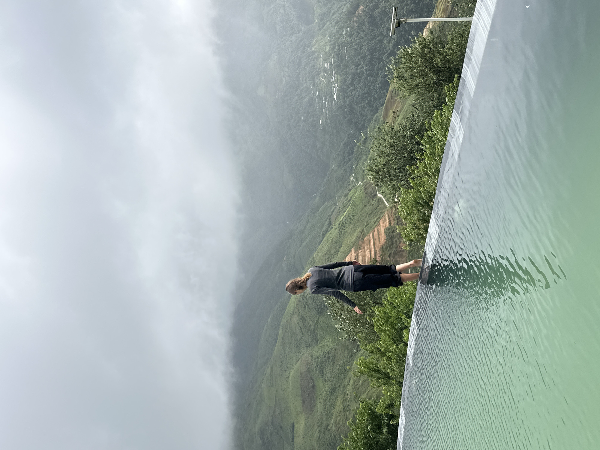 A woman stands at the edge of an infinity pool, looking out at a misty, mountainous landscape in Ta Xua, Vietnam. The pool's turquoise water reflects the overcast sky. The woman, facing away from the camera, wears a gray long-sleeved shirt and black pants. Her left foot is in the water. The background features lush green terraced hillsides and distant villages shrouded in fog. A solar-powered light fixture is visible to the right of the frame.