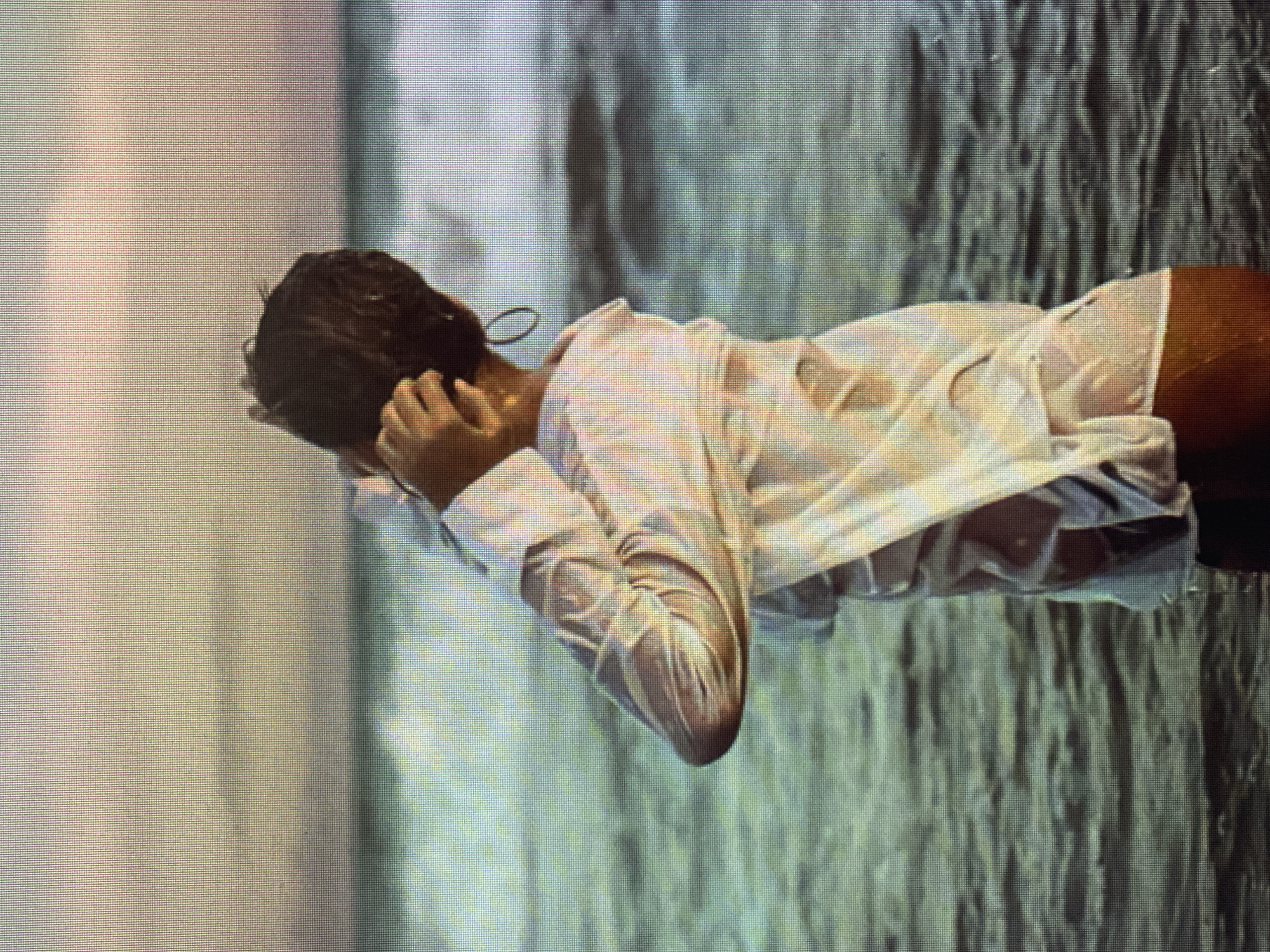 The media file depicts a close-up, vertical shot of a person lying down, partially submerged in water. The individual has dark hair and is wearing a white, wet shirt and orange bottoms. The shirt clings to the body, revealing the form beneath, and the fabric is translucent in places. The person's head is tilted down, and one hand is touching their head. The background is abstract, with vertical lines and ripples suggesting a textured surface, possibly water or a similar fluid. The lighting is soft and diffused, creating a serene and intimate atmosphere. The overall impression is one of quiet contemplation or relaxation, with the water and the wet clothing contributing to a sensual and evocative mood. The image is composed of what appears to be a pixelated display, indicative of a photograph taken of a screen or a low-resolution digital image.