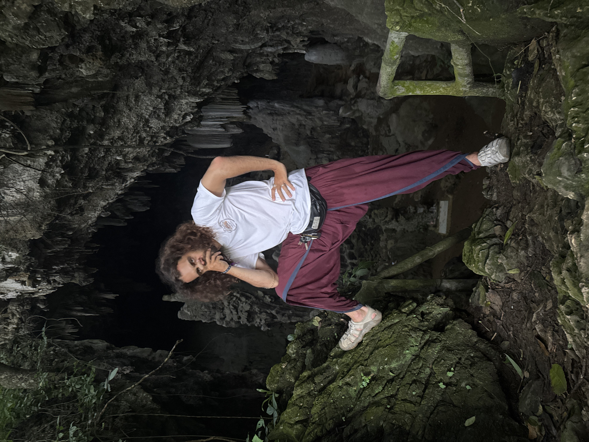 The image captures a man standing in the entrance of a cave in Mai Chau, Vietnam. The man has curly brown hair and is wearing a white t-shirt, maroon pants, and grey athletic shoes. He is posing with one hand on his chin and the other on his hip. The cave's interior is dark and features stalactites hanging from the ceiling and stalagmites rising from the ground. The foreground of the image shows moss-covered rocks and foliage, suggesting a natural, outdoor environment. A wooden railing is visible on the right side of the image, indicating a possible pathway or viewing area within the cave complex.