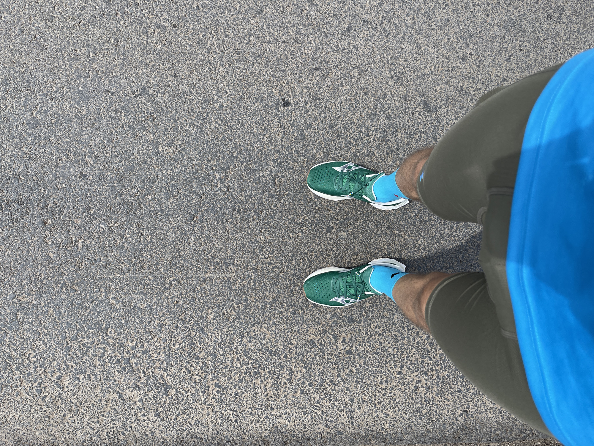 A first-person, eye-level shot depicts the lower portion of a person standing on a paved surface. The person is wearing green running shoes with white soles and light blue socks. The individual is also wearing olive-green shorts and a bright blue shirt is visible on the right side of the frame. The paved surface occupies the majority of the left side of the image. The location is not explicitly mentioned in the image; therefore, I cannot confirm it to be Перевозное, Russia.