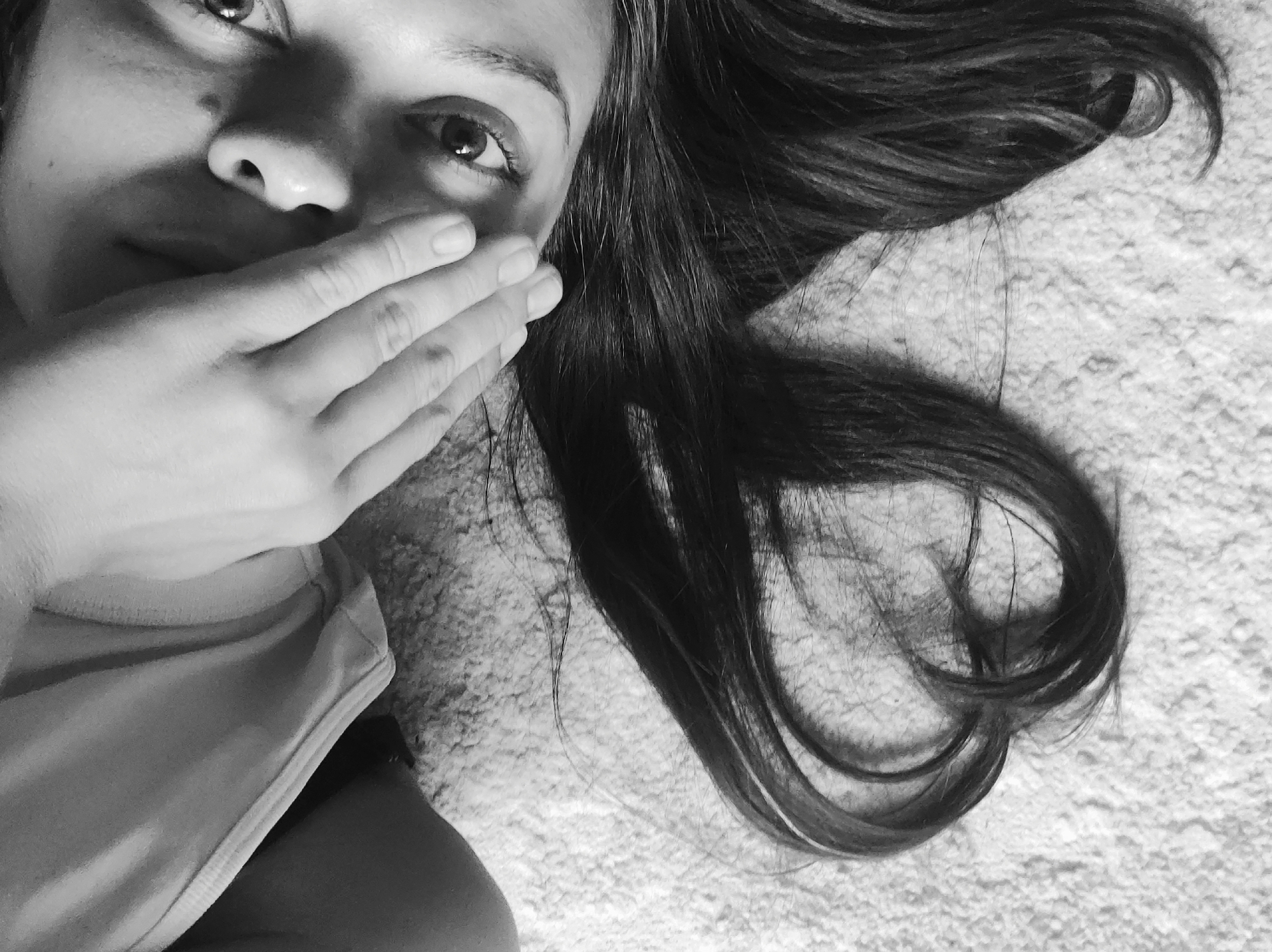 A black and white medium shot depicts a woman's face and hair against a textured background. The woman's face is partially obscured by her left hand, covering her mouth. Her eyes are visible and directed towards the viewer. Long, dark hair flows across the textured surface behind her head. She appears to be wearing a sleeveless shirt or tank top. There is no indication of the location in the city of Chaikovsky, Russia, or any specific environment. The scene appears indoors, possibly on a bed or carpet.