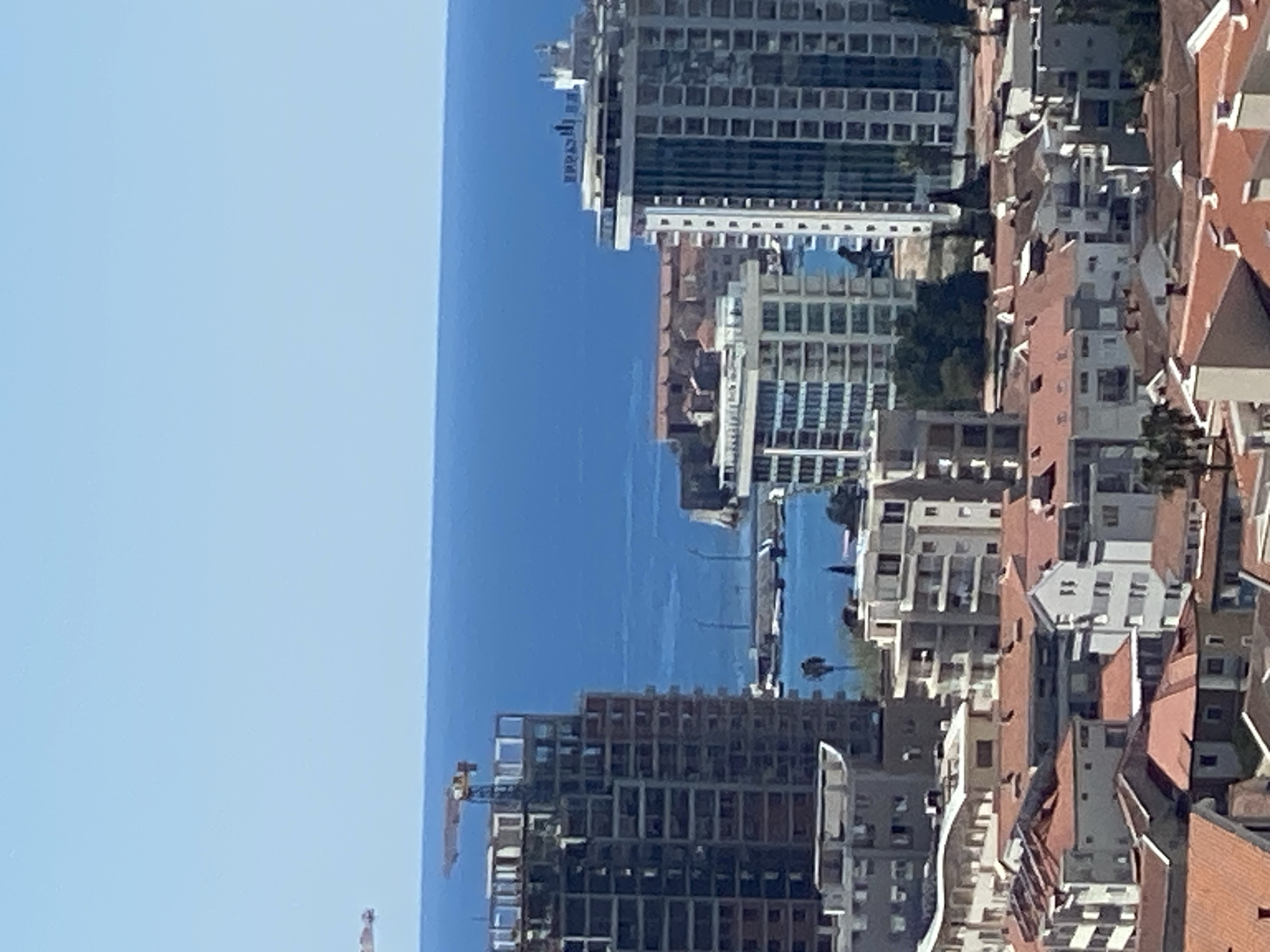 This aerial view features a coastal cityscape, likely Budva, Montenegro, with a clear, blue sky and body of water in the background. Buildings vary in style, with a mix of high-rise apartments and traditional red-roofed structures. A construction crane is visible near a high-rise under construction. There is minimal evidence of people or human activity in the frame.