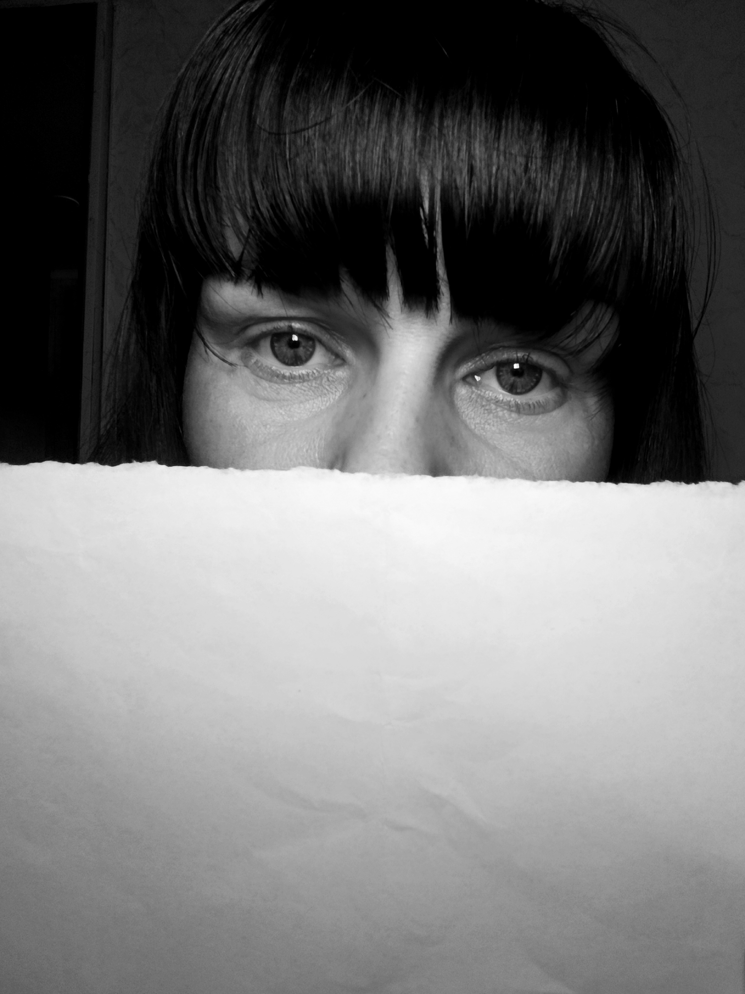A close-up, black and white photograph features a person with dark hair and bangs. The person's face is partially obscured by a white, rectangular piece of paper held directly in front of them, covering the lower half of their face. The focus is on their eyes. The background is dark and indistinct, appearing to be an indoor setting.