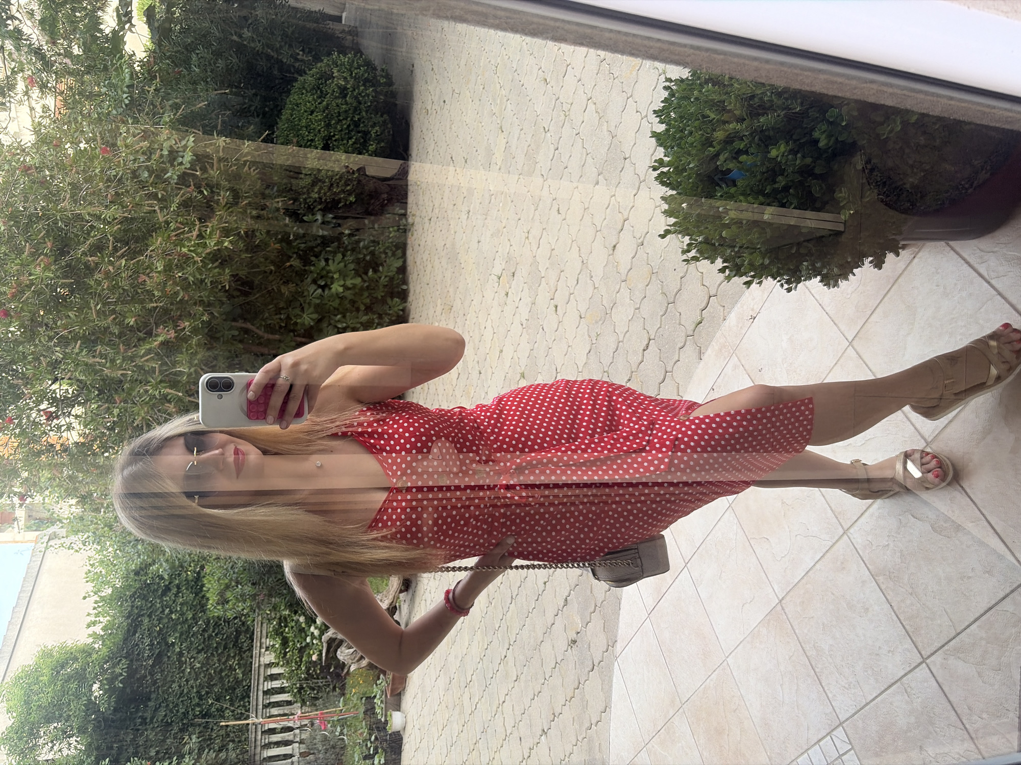 A young woman is taking a selfie using a phone in what appears to be a mirror or glass surface, which reflects an outdoor scene. She has long blonde hair and is wearing sunglasses and a red and white polka-dot dress. She is also wearing gold-colored sandals. She is holding a grey handbag with a chain strap. The reflection shows a paved outdoor area, greenery, and some buildings. The paving is light-colored and patterned. The greenery consists of bushes and trees. Based on the appearance of the environment, this photograph was most likely taken in Budva, Montenegro.