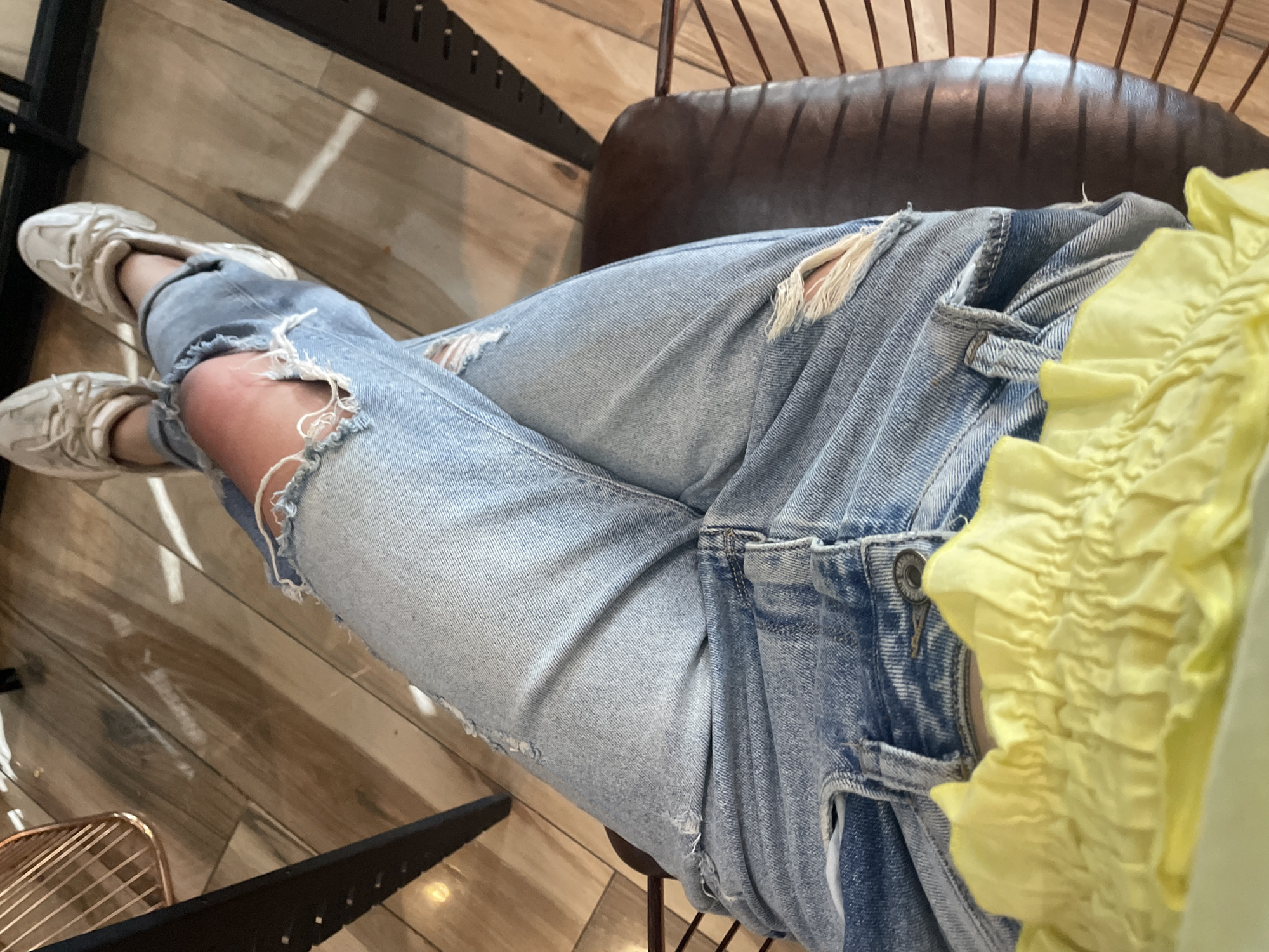A low-angle, close-up shot captures the lower body of a person sitting indoors. The primary subject is a pair of light-wash, distressed denim jeans with prominent rips at the knees, revealing skin underneath. The person is wearing white athletic sneakers with light-colored laces and soles. A portion of a bright yellow, ruffled or ruched top is visible on the right side of the frame, contrasting with the blue jeans.
The person is seated on a chair with a dark brown, possibly leather, back and metal frame details. The background consists of a light-colored wooden floor with horizontal planks, reflecting some ambient light. Parts of a metal chair base or leg are visible on the left and bottom left of the image. The person appears to be sitting casually, with legs crossed or resting comfortably. The setting suggests an indoor environment, possibly a cafe, restaurant, or lounge area, consistent with the provided location context of Sofia, Bulgaria. No specific time of day or visible text can be determined from the image.