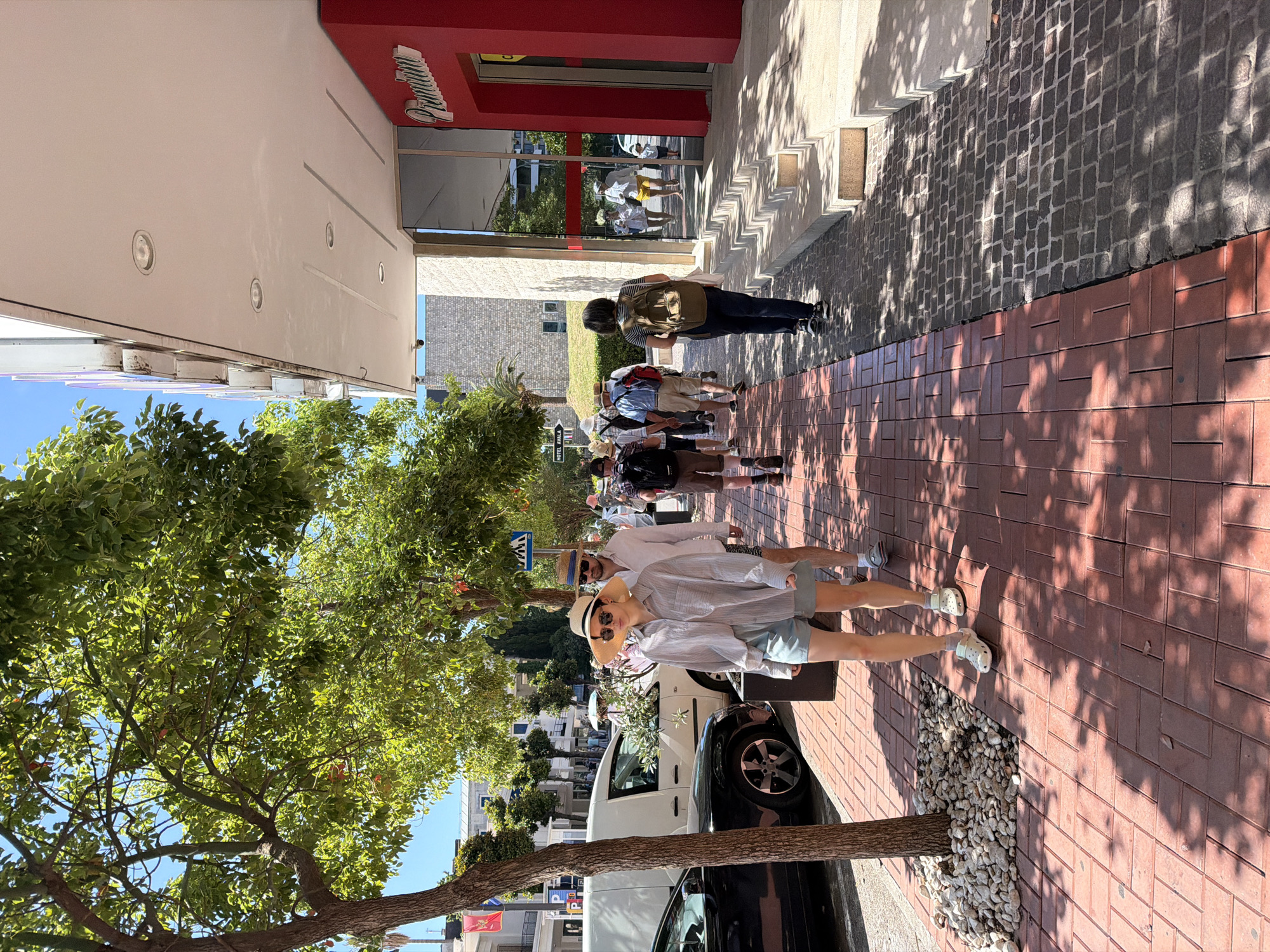 This image depicts a sunny urban street scene in Budva, Montenegro, identifiable by a visible "HOTEL AVALA" sign. The perspective is from a low angle on a sidewalk, looking down a street lined with trees and buildings. In the immediate foreground, a light-skinned woman walks on a red brick sidewalk, facing partially left. She wears a wide-brimmed straw hat, sunglasses, a light-colored long-sleeved button-down shirt, light blue shorts, and white Crocs-style sandals with white socks. A light-colored tote bag hangs from her left shoulder. Directly behind her and slightly to her right, a light-skinned man walks, wearing a straw hat, sunglasses, a light-colored long-sleeved shirt, and dark swim shorts. Further along the sidewalk, a person with dark hair, wearing a striped top and dark trousers, carries a light brown backpack and walks away from the viewer. Behind this individual, a group of approximately five to seven people, mostly men, are also walking away, many wearing hats and carrying backpacks, suggesting a tour group. One person in this group wears bright yellow shorts. The sidewalk itself is segmented. The left portion, adjacent to the street, is paved with reddish-brown rectangular bricks laid in a herringbone pattern. The right portion, closer to the buildings, is paved with dark grey cobblestones in a square pattern. Several mature trees with dense green foliage and visible reddish-orange fruits (likely citrus) line the sidewalk, casting strong, distinct shadows onto the pavement. Circular tree grates filled with white gravel surround the base of these trees. To the left of the sidewalk, a black car is partially parked, followed by a white van. Across the street, various light-colored buildings are visible, some displaying flags, including a red and white Montenegrin flag. A blue square sign with a white pedestrian crossing symbol is mounted on a pole above the middle-ground pedestrians. A "P" (parking) sign is partially visible to the left of the white van. To the right, a multi-story building features a prominent red lower facade with a white, stylized illuminated sign reading "Premium." Above this, the building has a glass and light-colored stone exterior. A reflective glass section of this building prominently displays reflections of people walking, including an individual in a white top and yellow shorts. A series of light-colored steps lead from the cobblestone sidewalk up to the entrance of this building, with sharp shadows defining their contours. An overhead canopy with recessed lighting extends