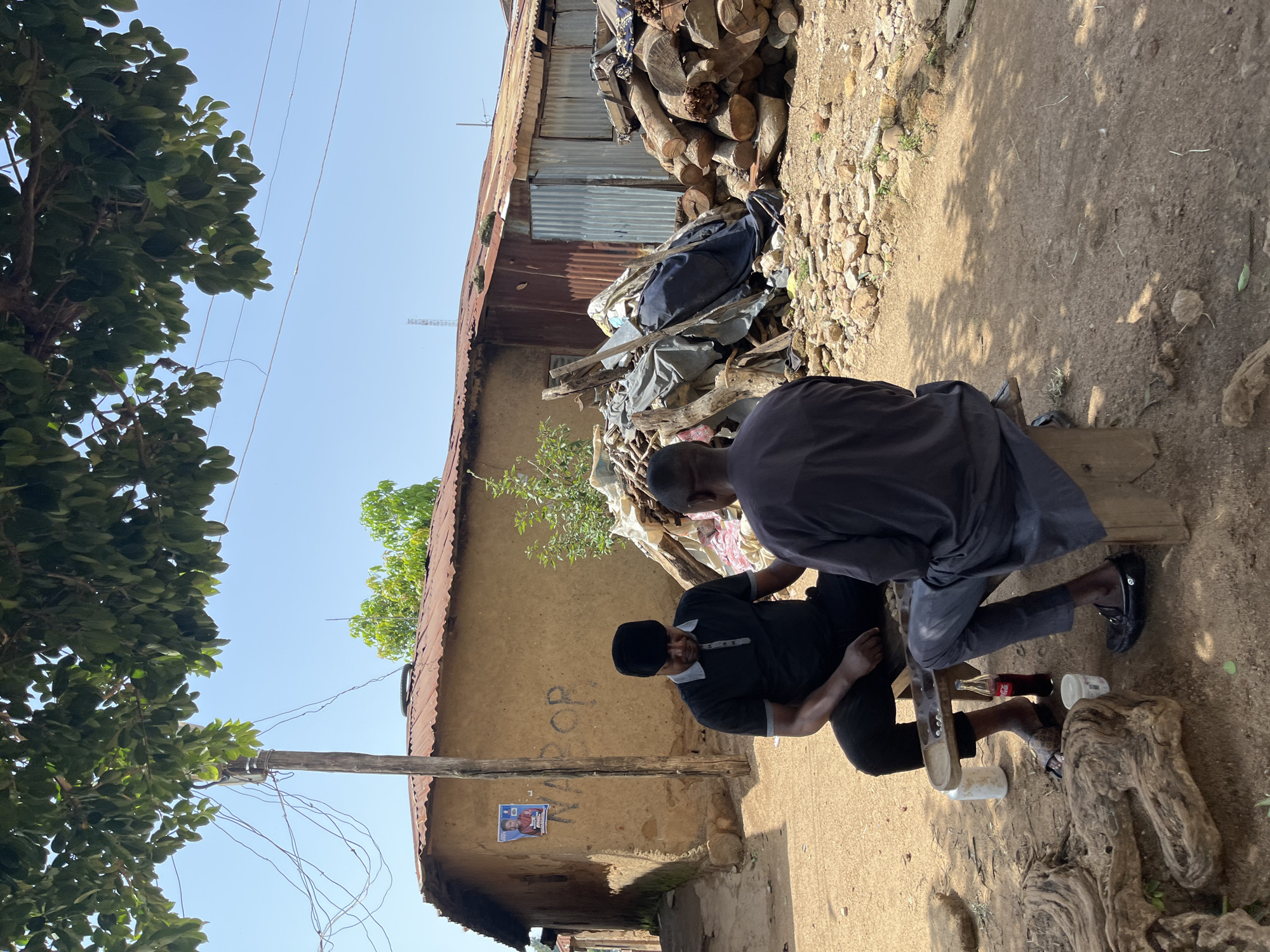 The image depicts an outdoor scene in a village setting, likely within Jos, Nigeria, on a sunny day. Two men are seated on a wooden bench playing a board game, possibly Ayo, indicated by the carved wooden board with multiple depressions filled with small white objects. The man on the left is wearing a black polo shirt and black trousers and a black cap, while the man on the right wears a grey, long-sleeved, loose-fitting garment. A bottle of Coca-Cola and a white cup are on the ground near the bench.
In the background, a single-story building with a corrugated iron roof is visible. The walls of the building appear to be made of earth or mud. A wooden pole with electrical wires is situated in front of the building. There's a poster of a man in a red shirt affixed to the wall, and the word "NABOR" is spray-painted on the wall below the poster. To the right of the building, a pile of stacked logs and some covered materials are visible. The ground is covered in dirt and small stones, with scattered debris. The overall environment suggests a rural or informal settlement area.