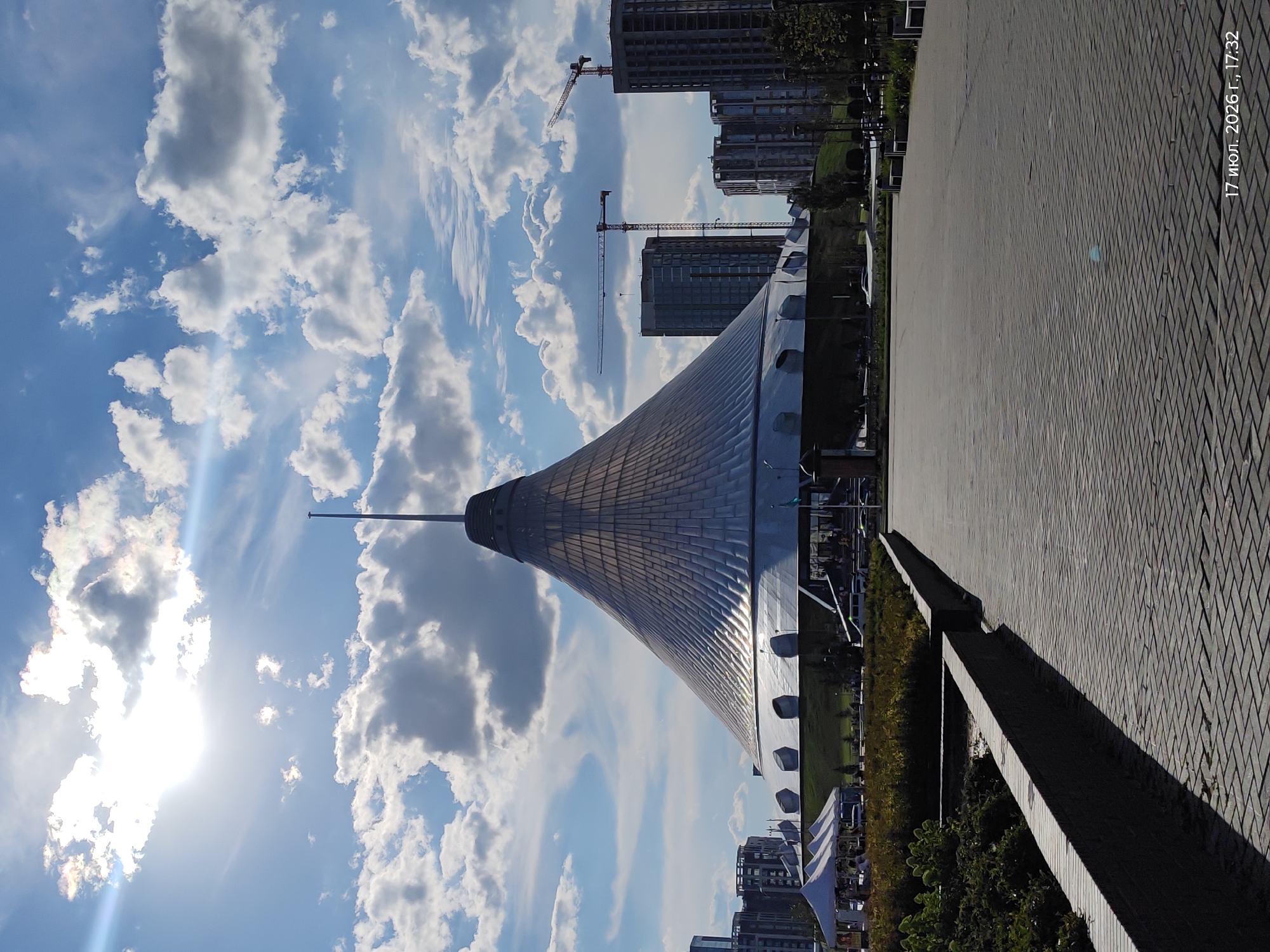 This is a landscape image taken outdoors on a sunny day with scattered clouds. The primary subject is a large, conical-shaped building with a metallic, tiled roof, which appears to be a modern architectural structure. In the background, several tall, modern buildings are under construction, indicated by the presence of construction cranes. The foreground shows a paved walkway with a brick-like pattern, and a raised concrete planter bed with greenery.
The time of day is indicated by the bright sunlight and the length of the shadows, suggesting it is late afternoon. The sky is a vibrant blue, with dramatic white and grey clouds, some of which are catching the sunlight, creating a somewhat ethereal effect. The overall atmosphere is one of urban development and a pleasant, sunny day.
The image includes text in the upper right corner: "17 июл. 2026 г., 17:32". This text indicates the date and time the photo was taken: July 17, 2026, at 5:32 PM. The location context of Astana, Kazakhstan, further helps to identify the architectural style and urban setting. No people are visible in the frame.