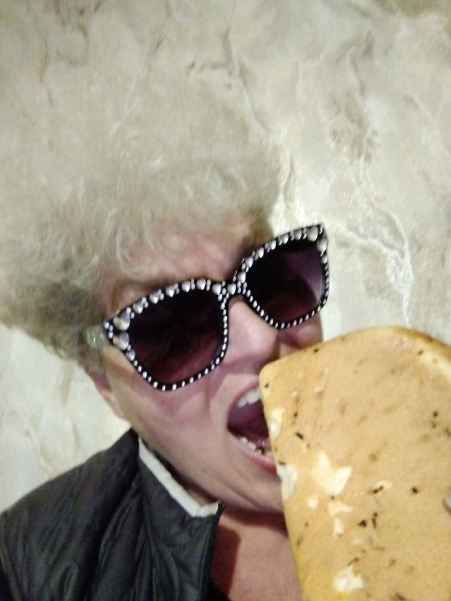 The image displays a close-up, slightly tilted portrait of an individual in the act of eating. The subject, likely an older person, has voluminous, light-colored (possibly white or very light blonde) hair. They are wearing prominent dark sunglasses with black frames, heavily embellished with numerous small, reflective, heart-shaped or rounded studs along the top and side edges.
The person's mouth is open wide, positioned to take a bite from a large, irregularly shaped piece of food. This food item appears to be a light brown or beige flatbread, possibly focaccia or a similar type, characterized by visible darker specks, likely herbs or spices, and lighter, uneven patches across its surface.
Part of a dark, possibly black, quilted or puffer-style jacket is visible on the left side of the frame, with a lighter-colored collar or shirt underneath. The background is a light-toned, textured surface, possibly a wall made of stone or patterned plaster, showing subtle variations in color and texture.
The image captures a candid moment of consumption, with the subject fully engaged with the food. No specific time of day or weather conditions can be determined. The scene appears to be indoors or against a wall. There is no visible text. The location context provided is Horishni Plavni, Ukraine, but the image itself offers no specific geographical cues beyond the background texture.