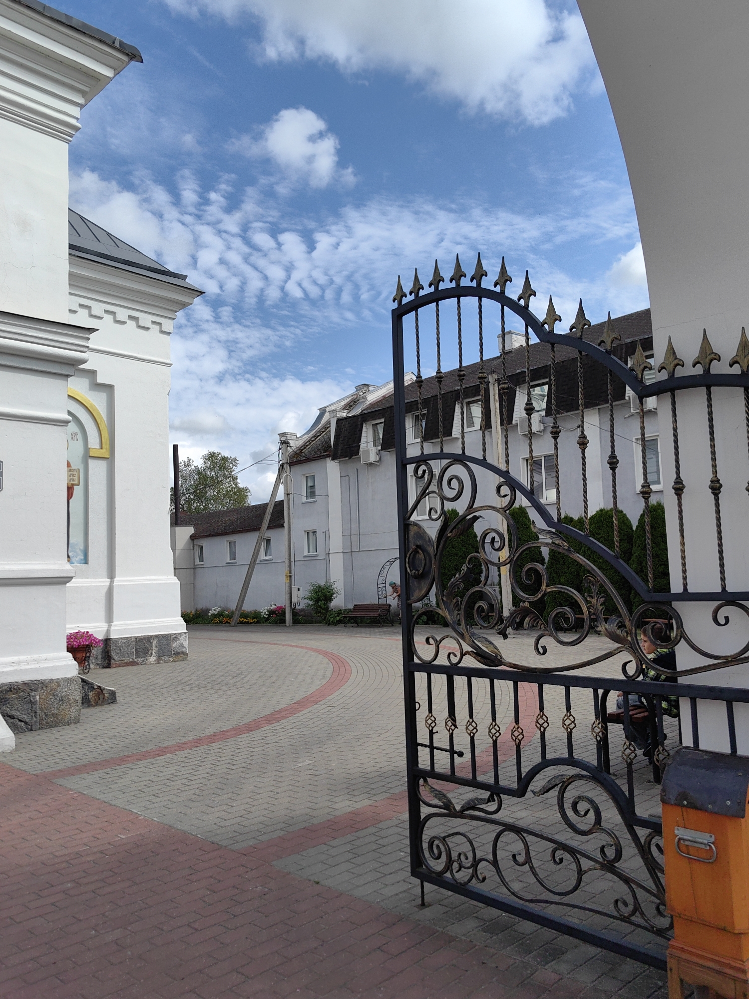 The image depicts an ornate black metal gate, partially open, in front of a white building with a grey tiled roof. The gate features decorative scrollwork and pointed finials. Beyond the gate, a light-colored building with multiple windows and dark sloped roofs is visible. The ground in the foreground is paved with interlocking bricks in shades of grey and red, with a red brick border forming a curved path. To the left, a portion of a white structure, possibly a church or monastery, is visible with decorative architectural details. A potted pink flower arrangement is placed near the base of this structure. In the background, a thin, tall structure, possibly a chimney or utility pole, rises above the buildings. The sky is blue with scattered white, wispy clouds. The scene appears to be an outdoor courtyard or entrance. A person is visible sitting on a bench behind the gate. An orange mailbox with a metal handle is situated to the right of the gate.