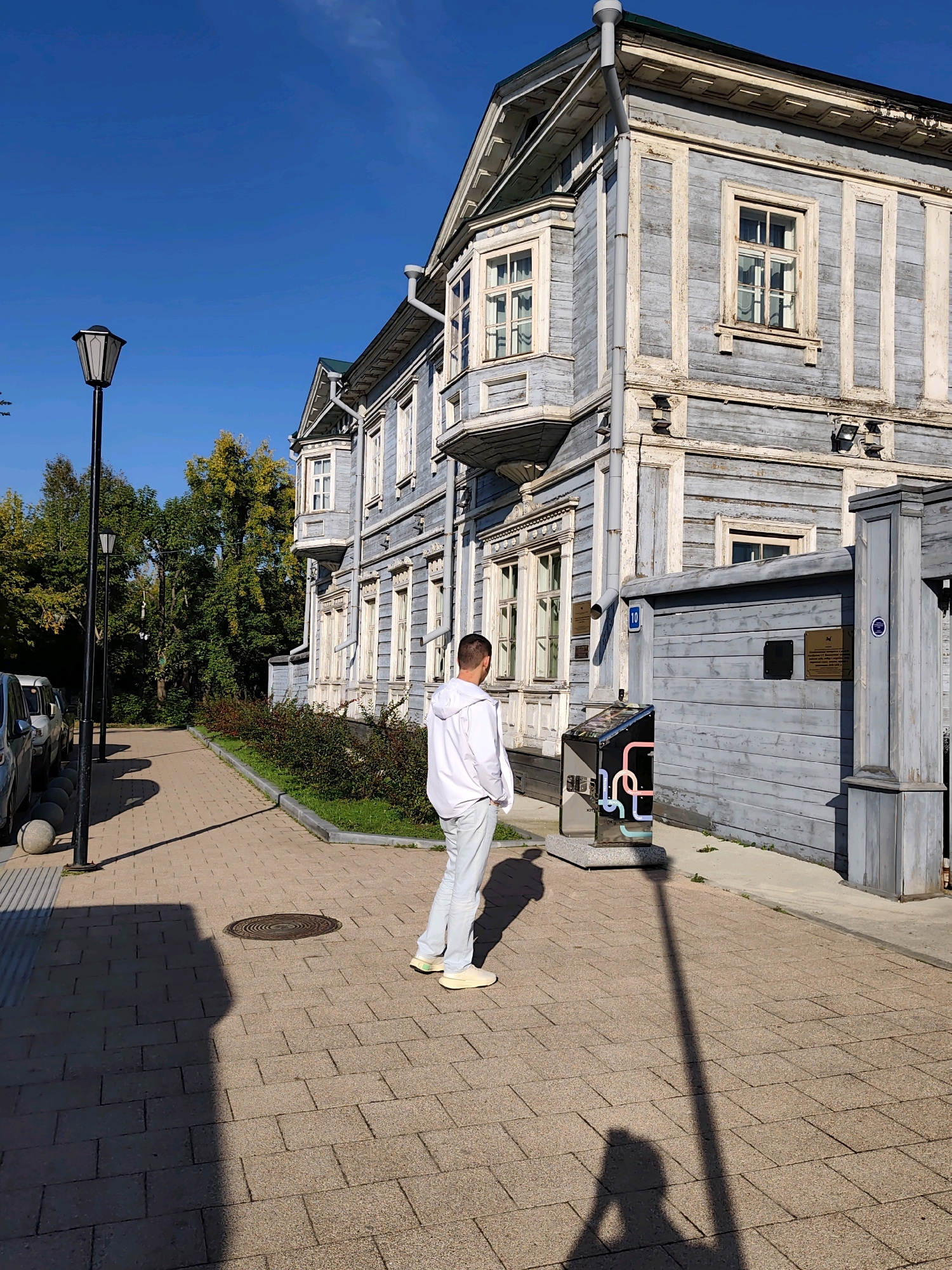 The image shows a person standing on a paved sidewalk in front of a historical wooden building with grey siding and white trim. The building has multiple windows and a bay window. To the left of the building, there is a black lamppost and several parked cars. Behind the building, there are lush green trees. The person is wearing a white hooded sweatshirt and light-colored pants, with their back to the camera. They appear to be looking at the building. In the foreground, there is a circular manhole cover on the sidewalk. A shadow of the person and the lamppost is cast on the ground. This scene is located at the Museum of the City of Irkutsk in Irkutsk, Russia.