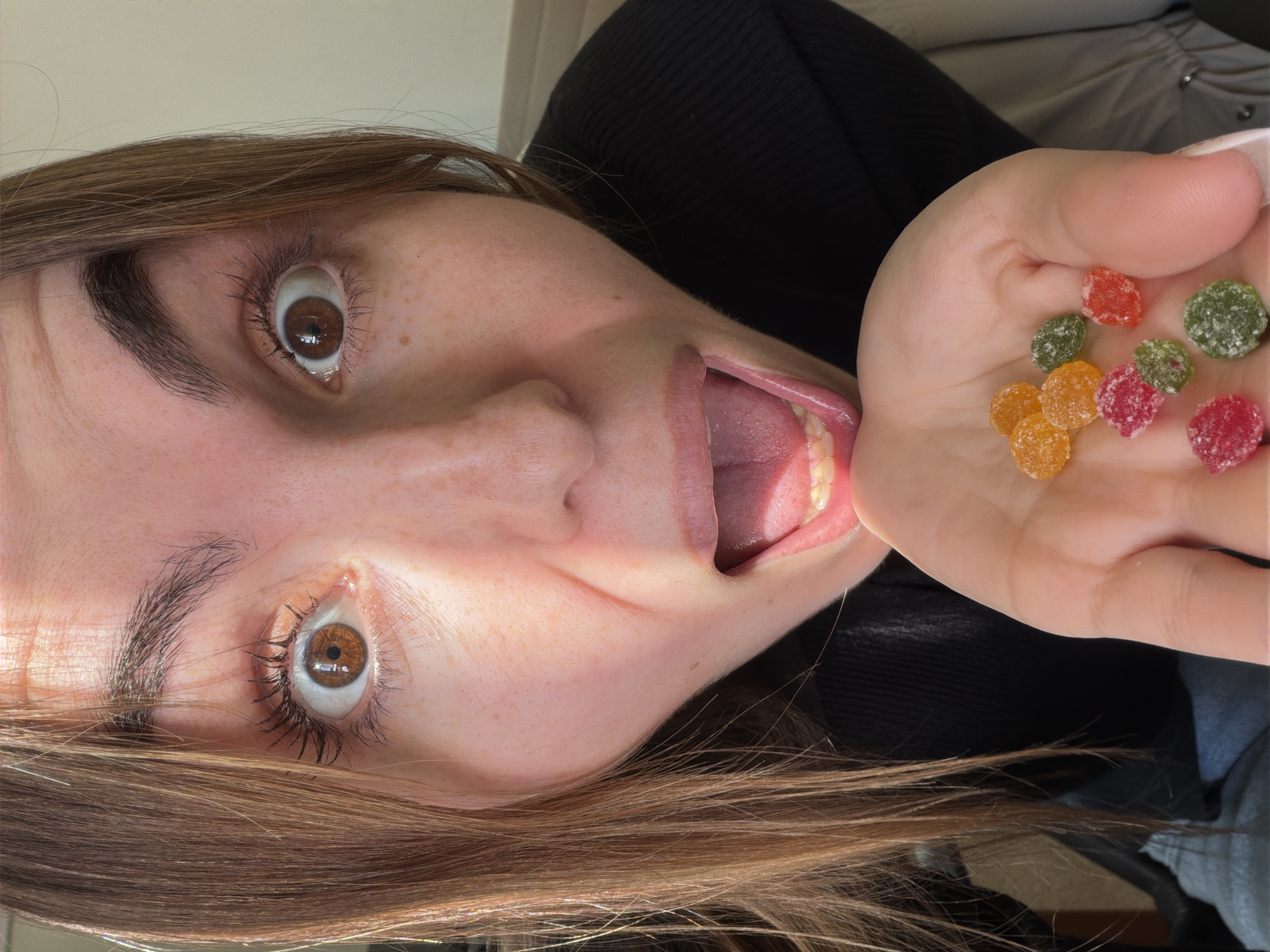 A person with light brown hair and brown eyes is lying on their back, holding a handful of multi-colored, sugar-coated gummy candies towards their open mouth. The person's eyes are wide, and they have freckles on their face. They are wearing a black top. The background is a muted grey color, possibly a couch or chair. There is no indication that this image was taken in Izhevsk, Russia.